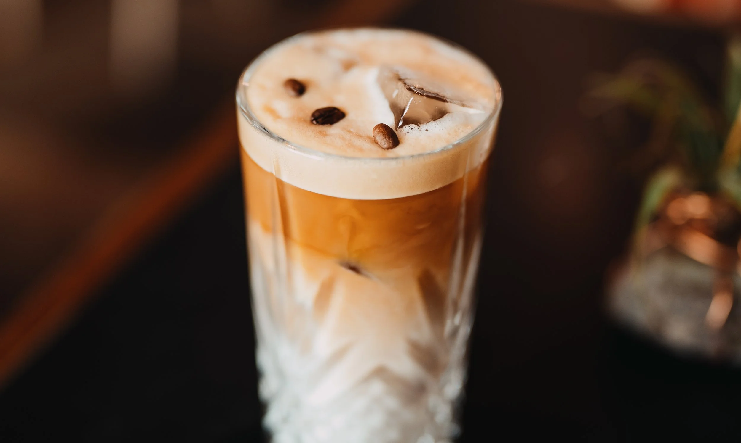 A tall glass of iced coffee with a frothy top, coffee beans, and ice cubes, on a dark surface.