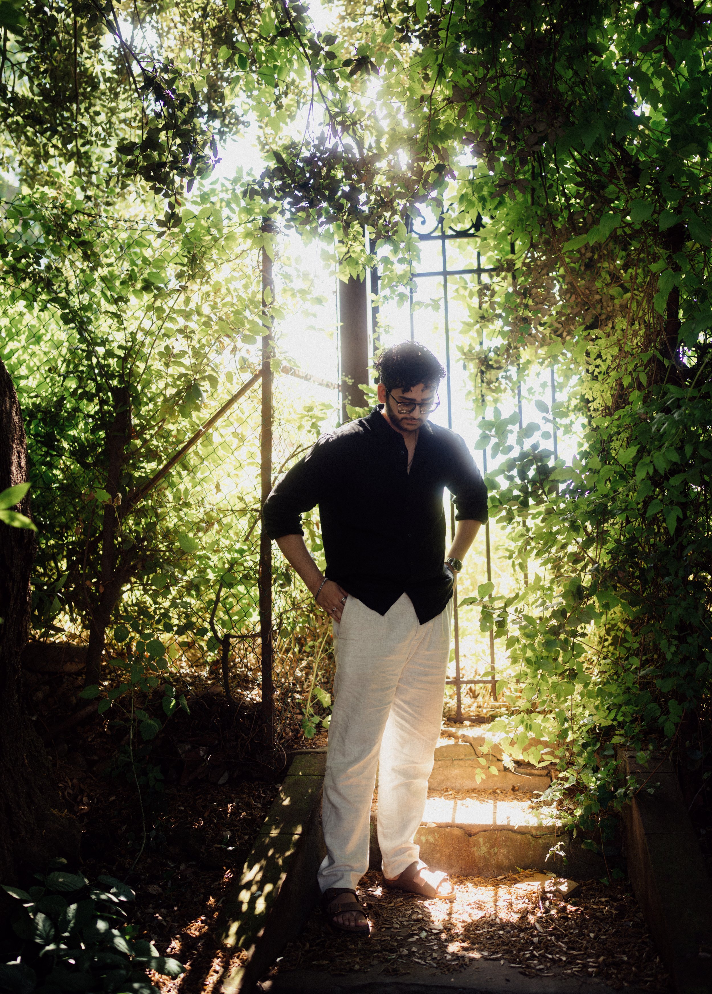 Man standing outdoors in a lush, green garden, looking down with hands in pockets, backlit by sunlight through trees and a metal gate.