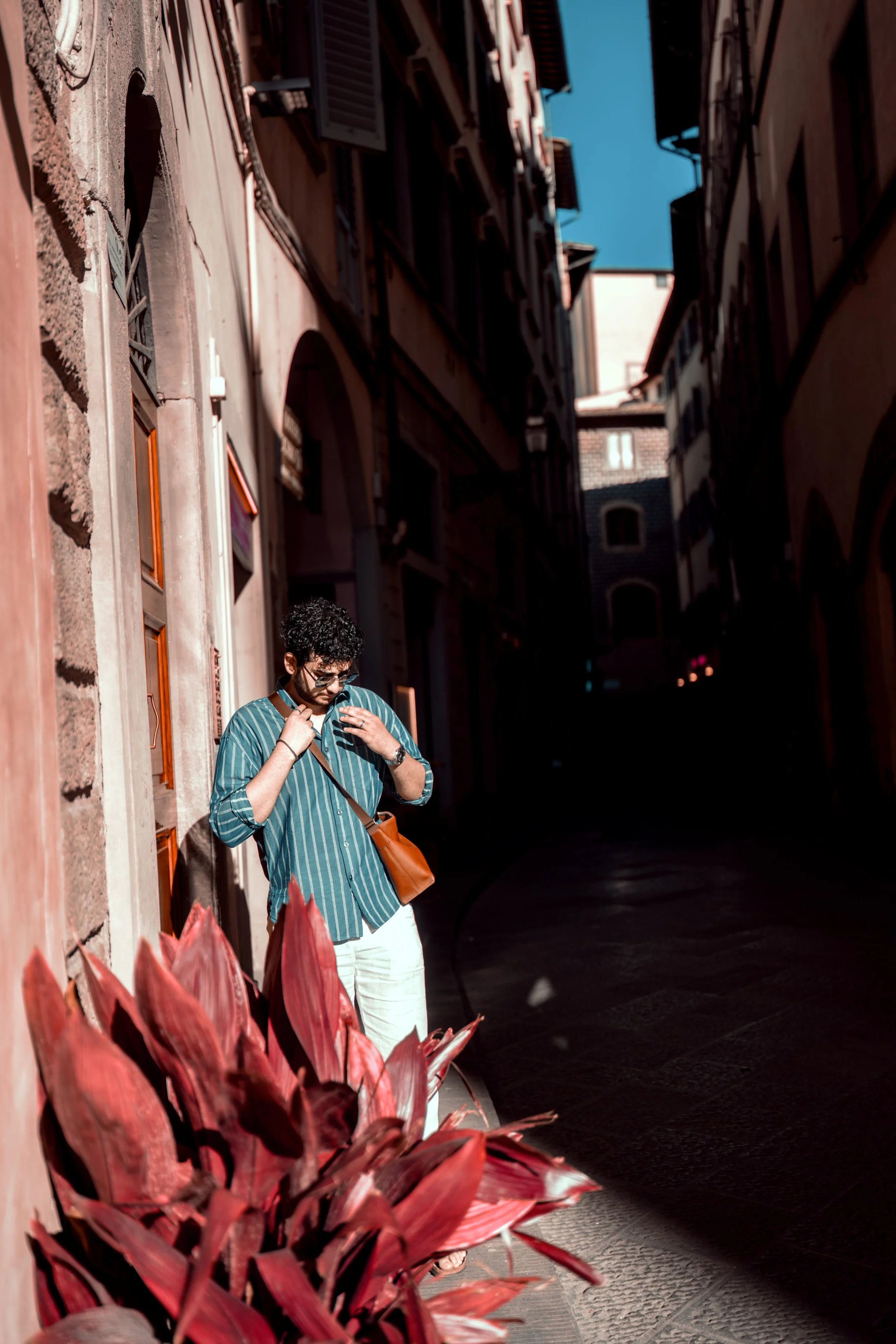 A man with curly hair, wearing sunglasses, a striped shirt, and light-colored pants, stands on a narrow city alleyway near red plants, with tall buildings on either side and a clear blue sky overhead.