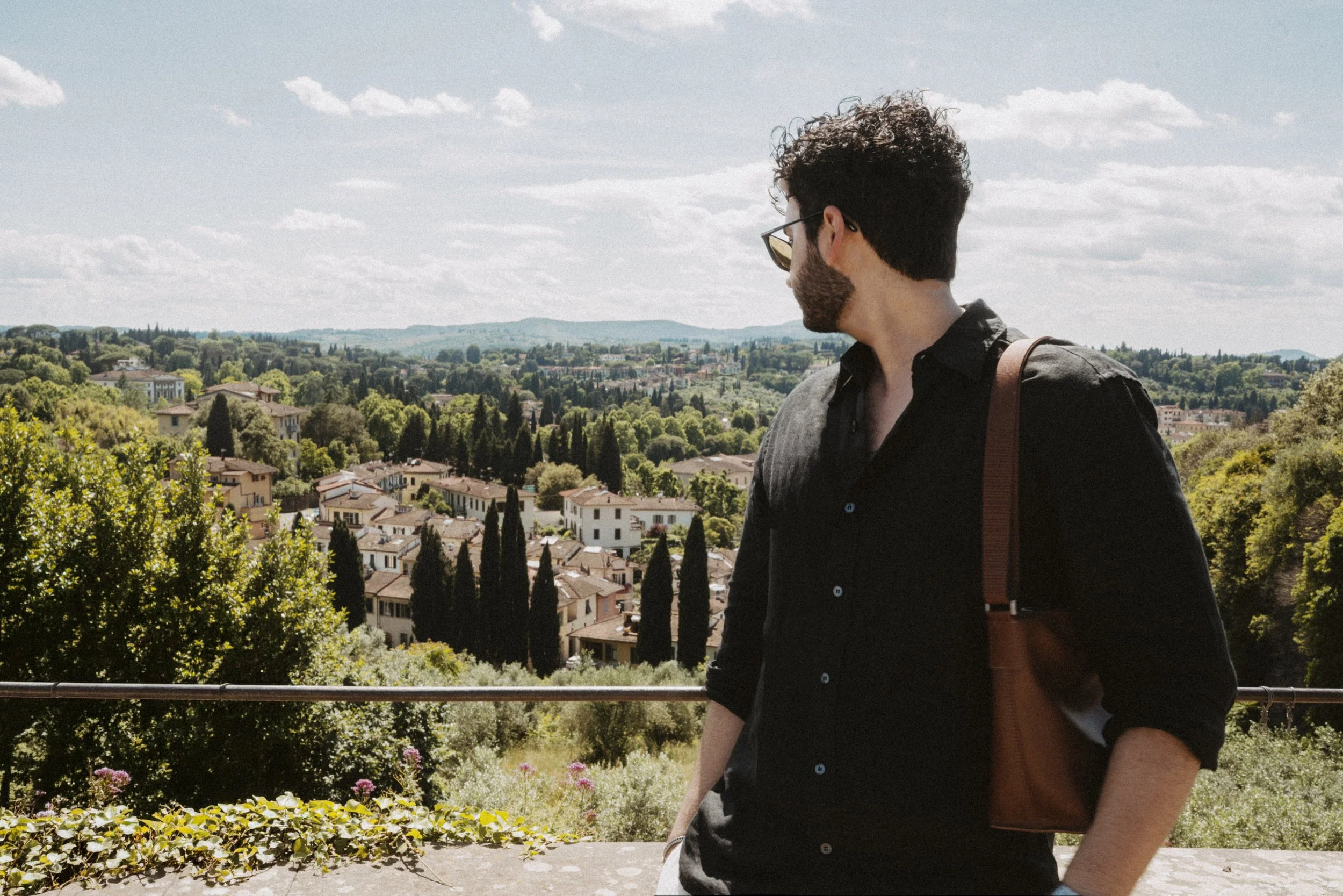 A man with curly dark hair, beard, wearing sunglasses, and a black shirt stands outdoors, looking to the right, with a scenic view of a town and green trees in the background on a partly cloudy day.