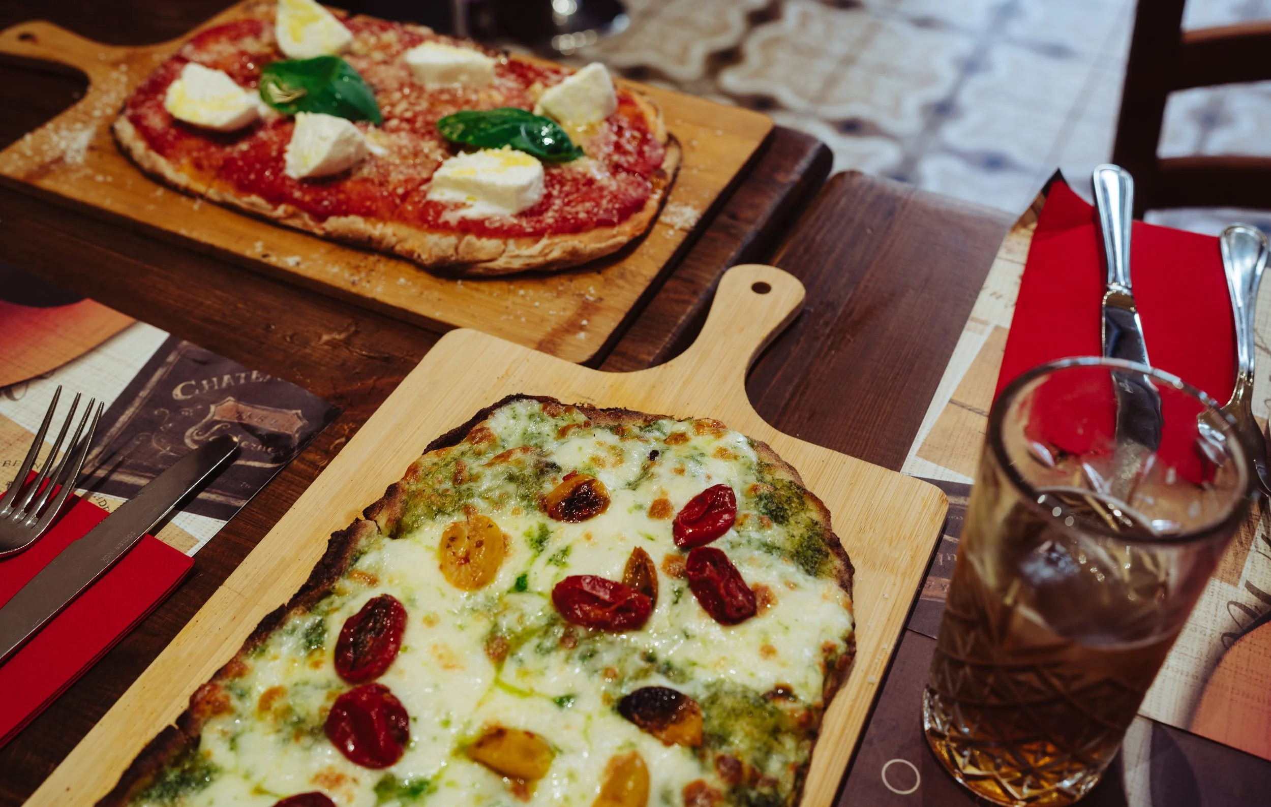 Two pizzas on wooden boards on a table, one topped with cheese and red and yellow cherry tomatoes, the other with cheese, tomato sauce, mozzarella, and basil leaves, next to a glass of dark soda with ice, utensils, a red napkin, and a paper placemat.
