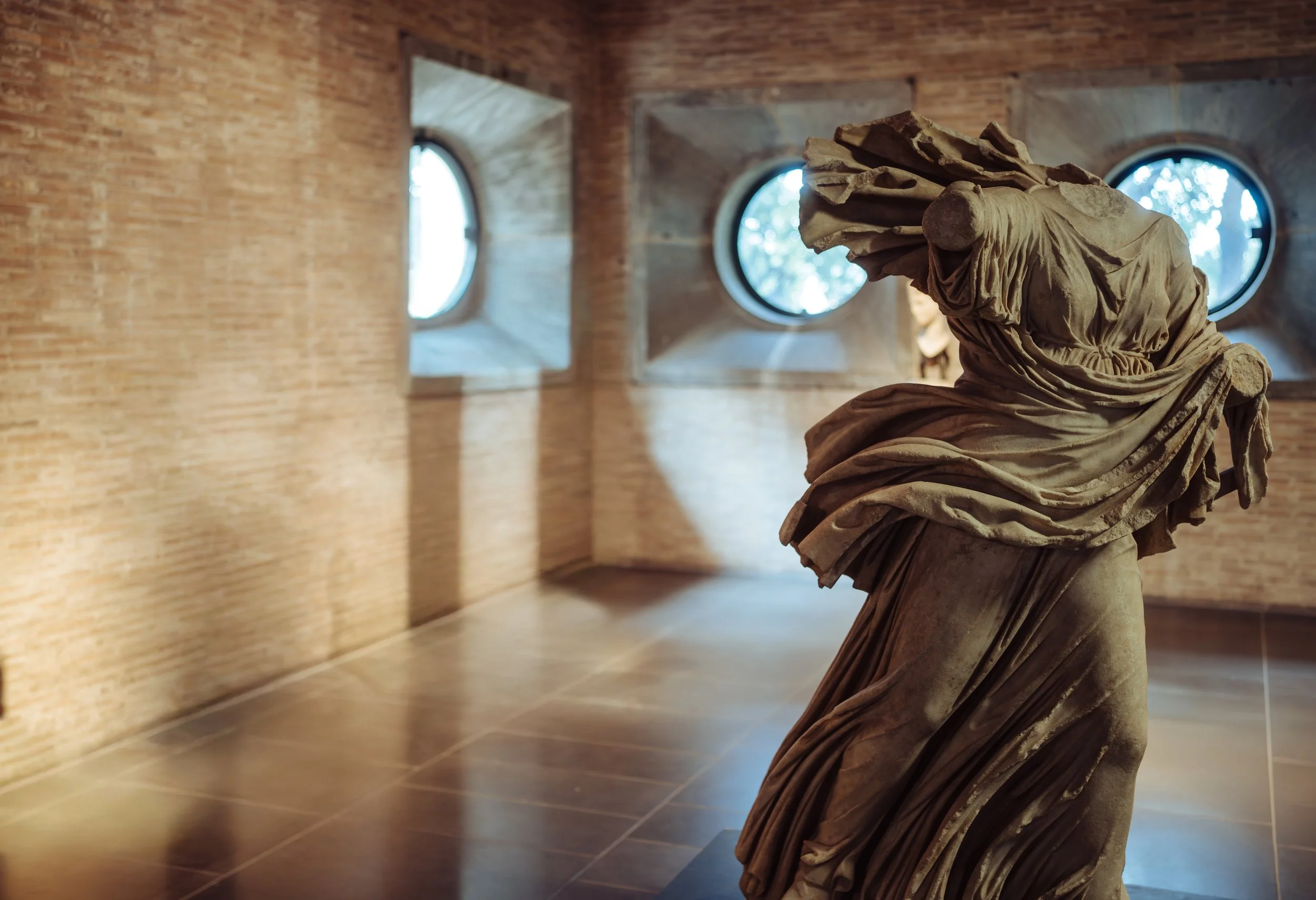 Statue of a headless woman in flowing robes inside a museum with brick walls and round windows.