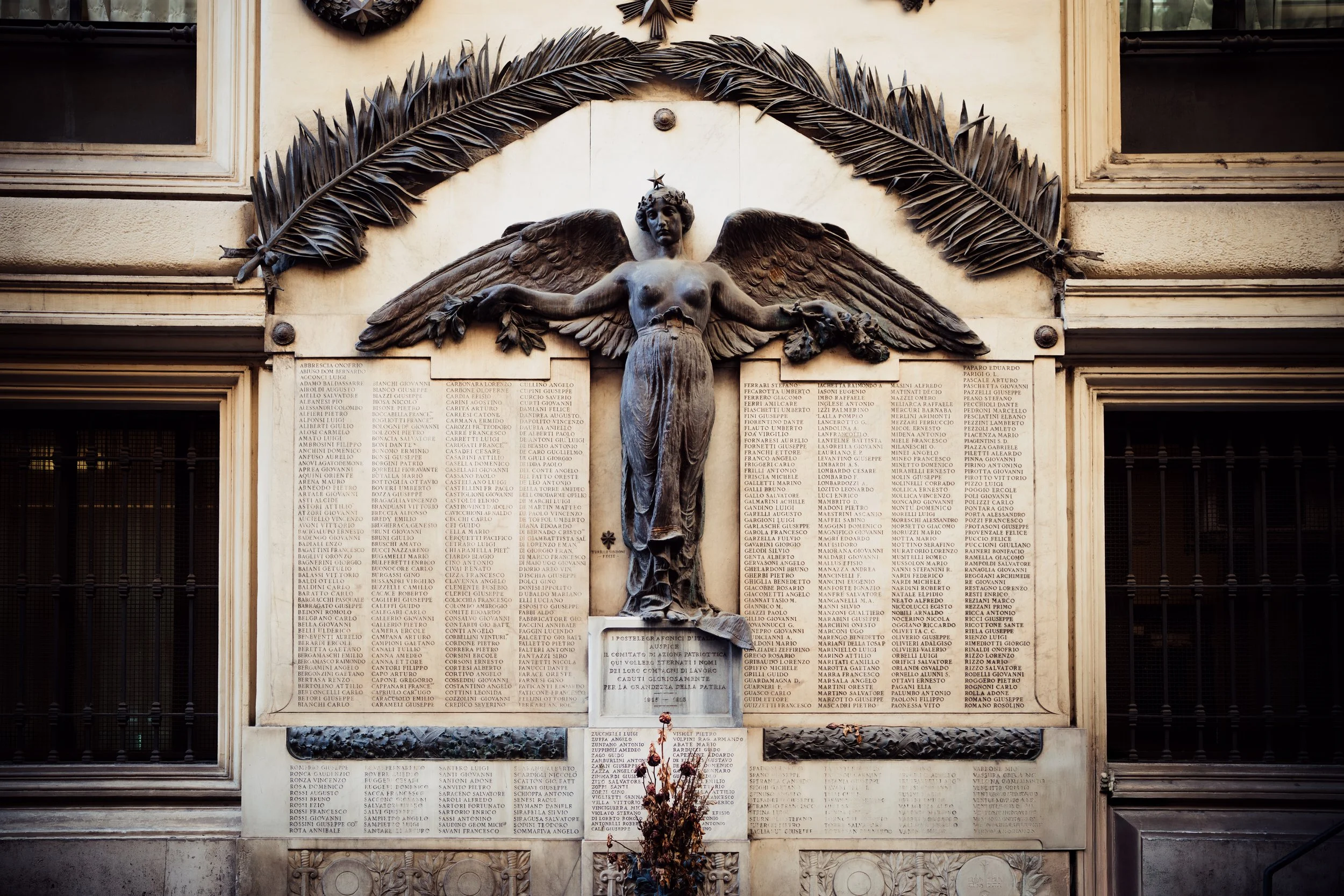 A memorial plaque featuring a bronze sculpture of a winged female figure with her arms outstretched, surrounded by columns of engraved names on a wall. There are two large decorative leaves above the sculpture.