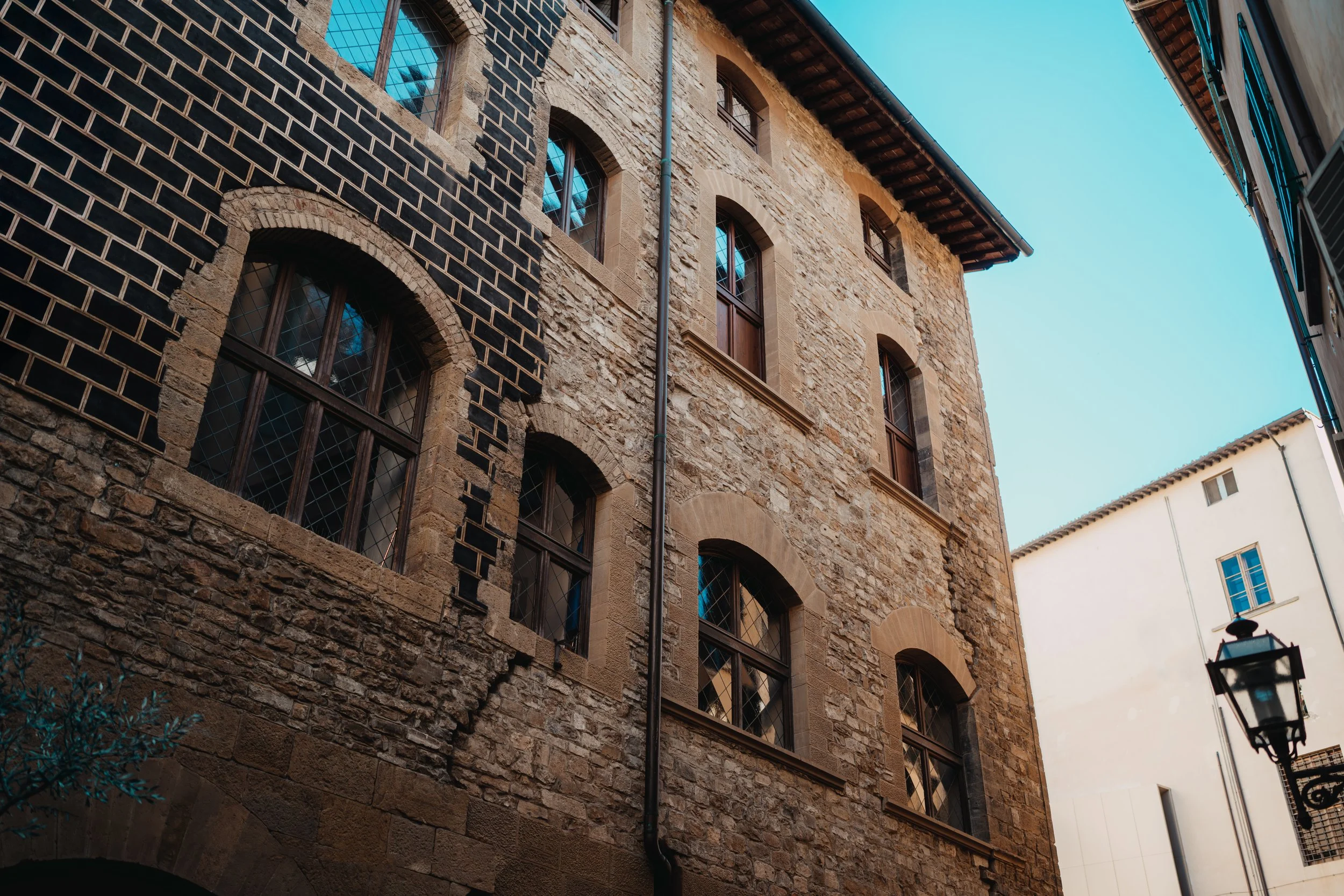 A tall stone building with arched windows and a mix of brick and stone exterior, located in a narrow alley with neighboring buildings, under a clear blue sky.