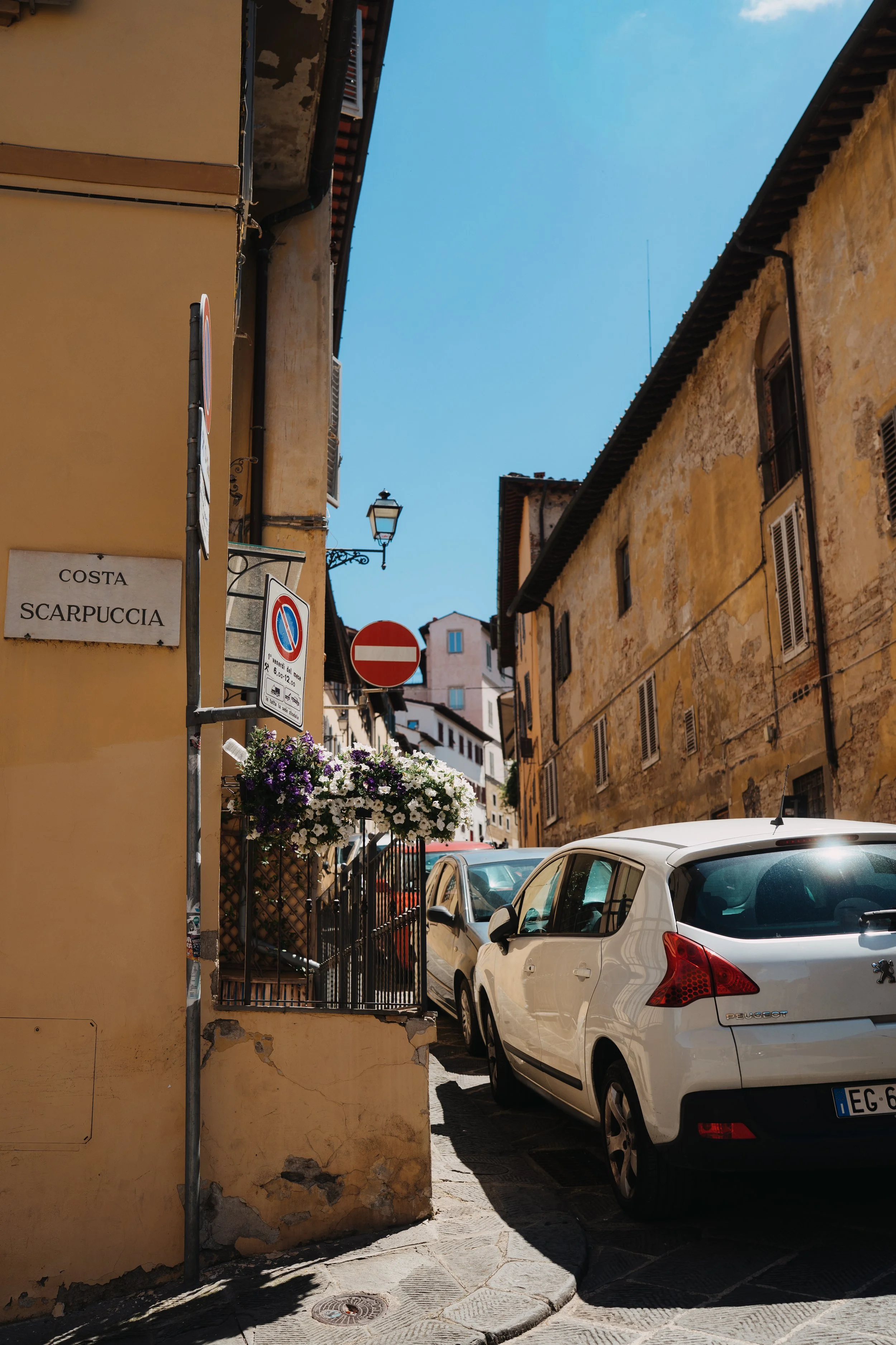 A narrow street in Italy with parked cars, old yellow buildings, and a blue sky.