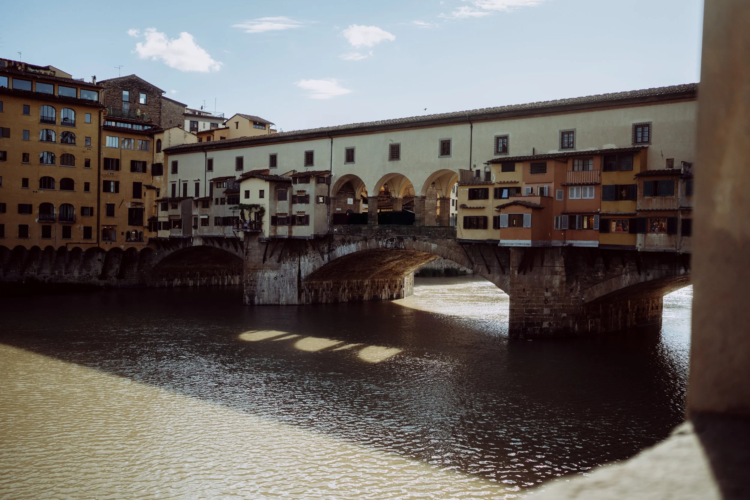 A bridge with multiple arches crossing a river, with buildings on either side of the bridge and a partly cloudy sky overhead.