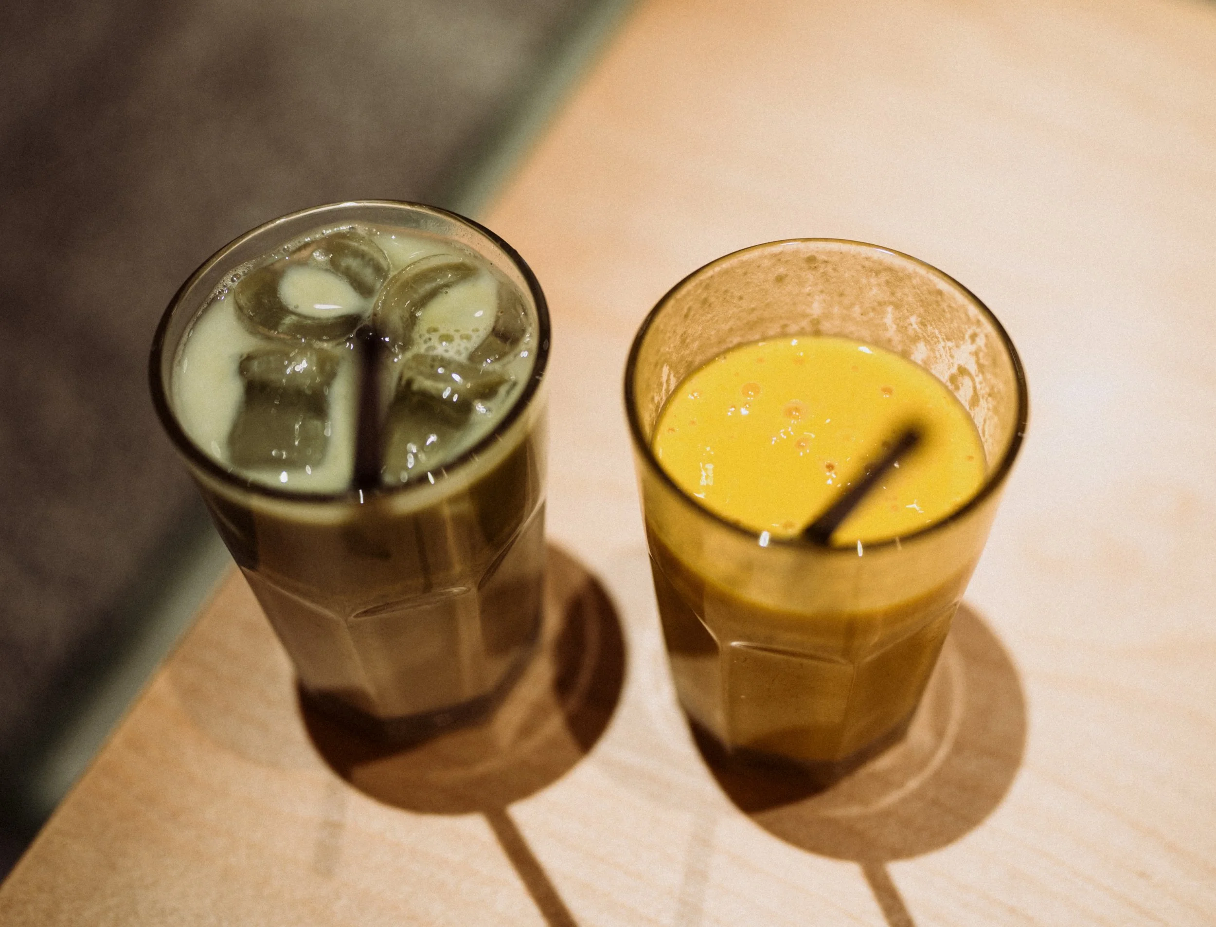 Two tall glasses on a wooden surface, one filled with iced coffee and the other with what appears to be orange juice, both with black straws, casting shadows.