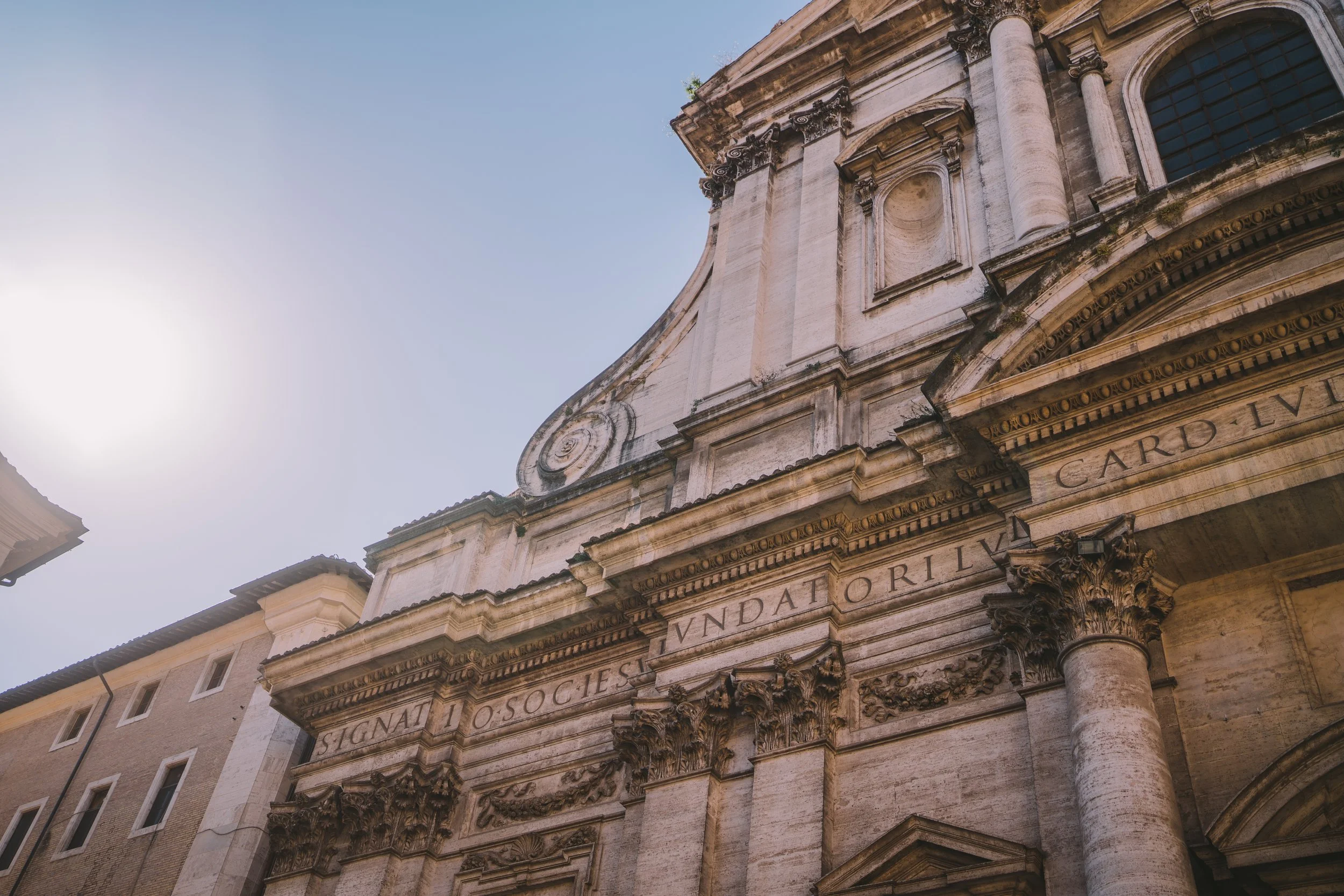 Close-up of an ornate, historic stone building with Latin inscriptions on its facade, columns, and decorative architectural details, set against a clear sky with the sun shining brightly.