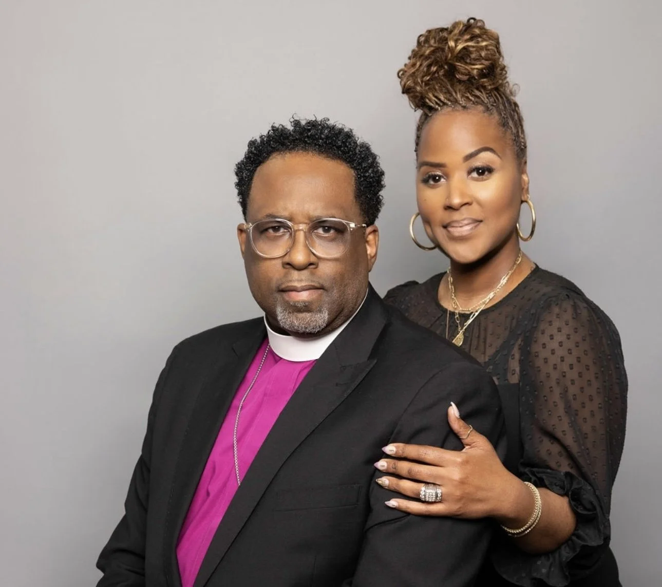 Two people pose together, one in a black suit with a magenta clerical shirt and the other in a black dotted blouse with an updo hairstyle.