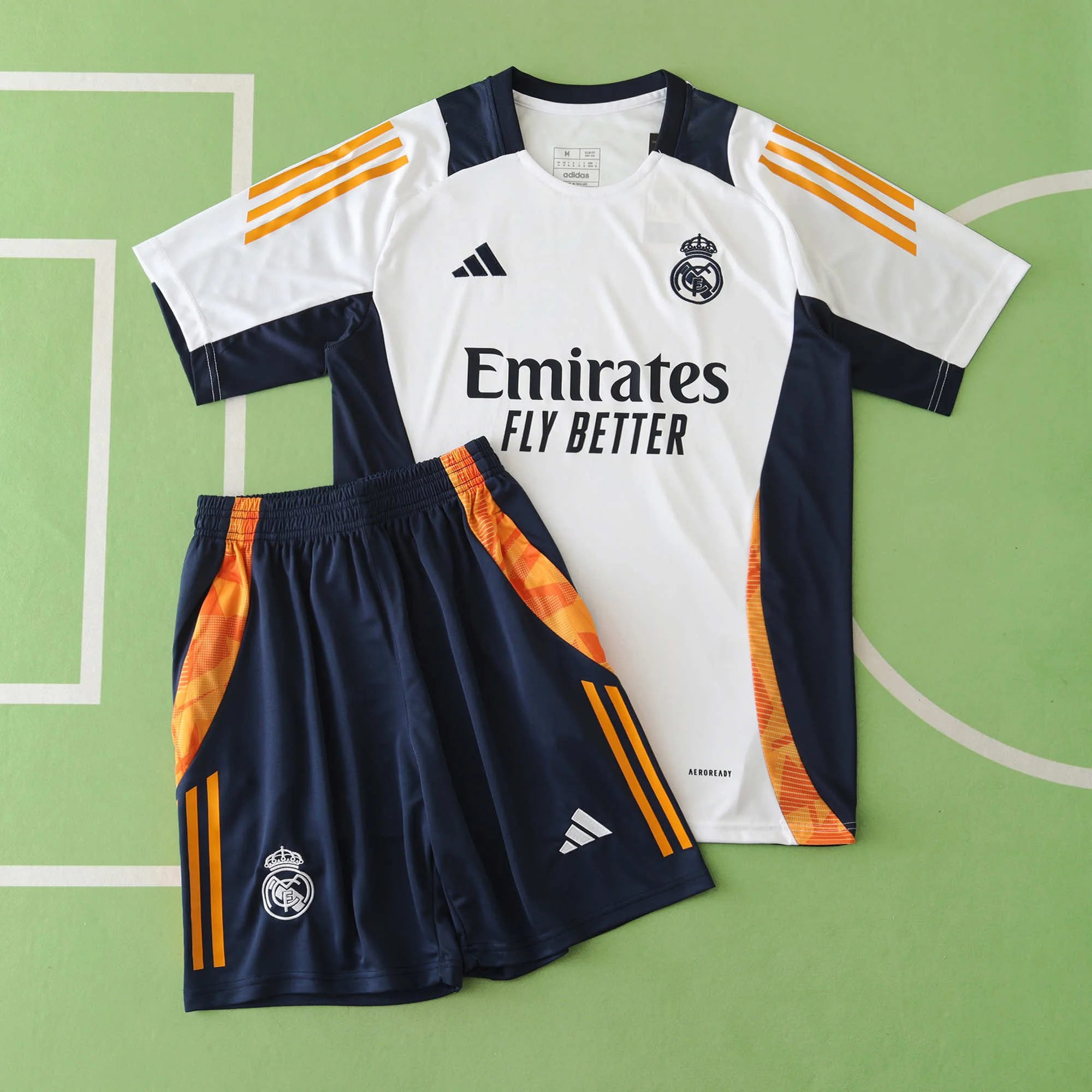 Real Madrid soccer uniform set includes a white and navy jersey with Adidas and Real Madrid logos, and matching navy shorts with orange accents, on a green sports court background.