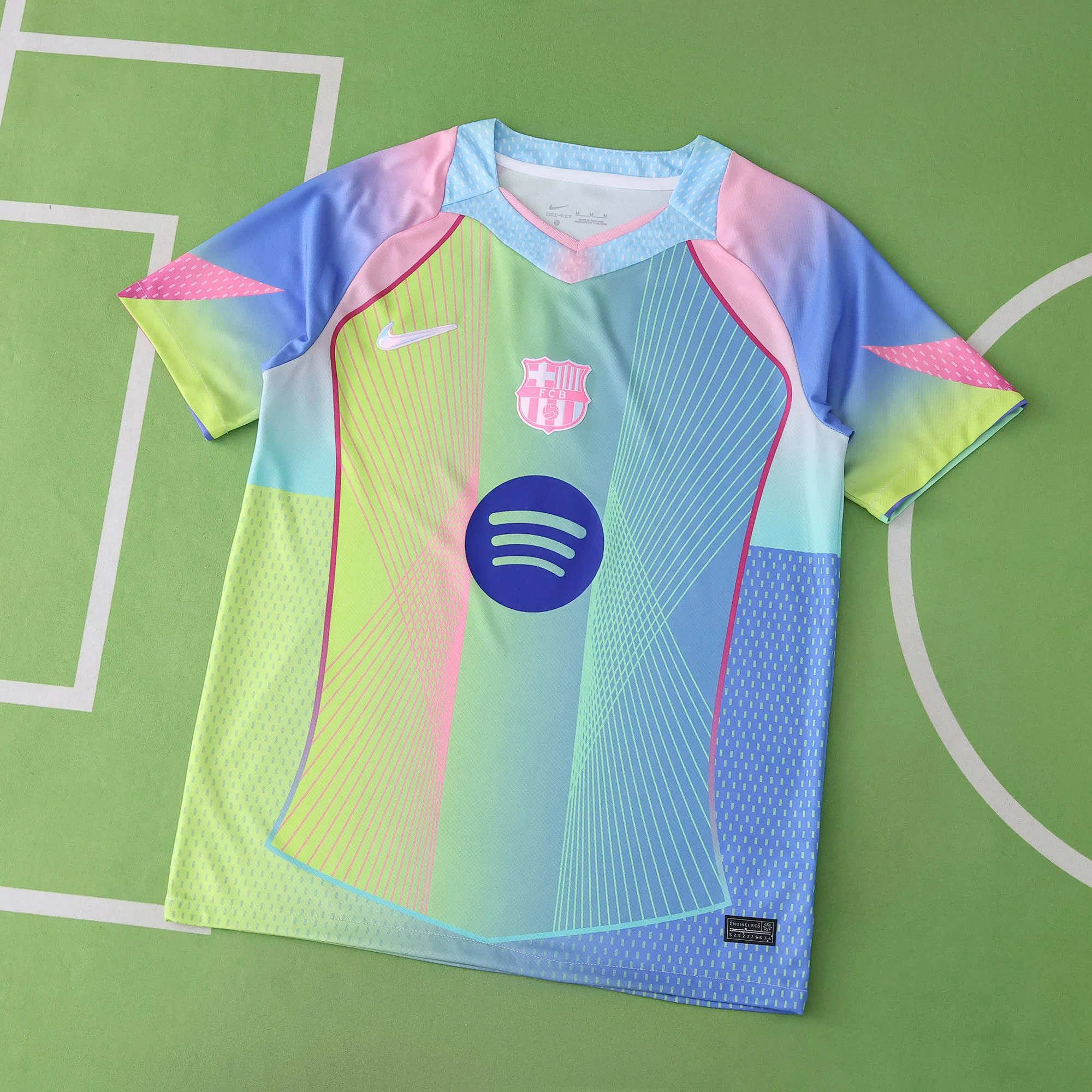 A colorful soccer jersey with pastel shades of pink, blue, green, and yellow, featuring the FC Barcelona crest, Spotify logo, Nike logo, and a size tag, laid flat on a green surface with white lines.