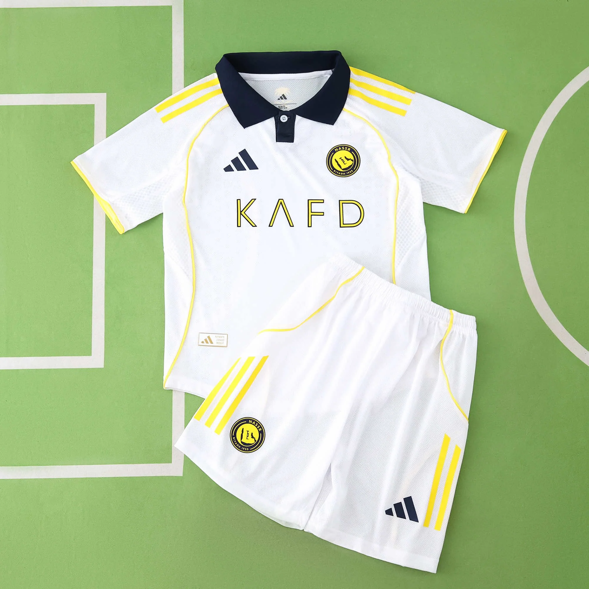 White soccer jersey and shorts with yellow and black accents on green background, displaying logos and text.