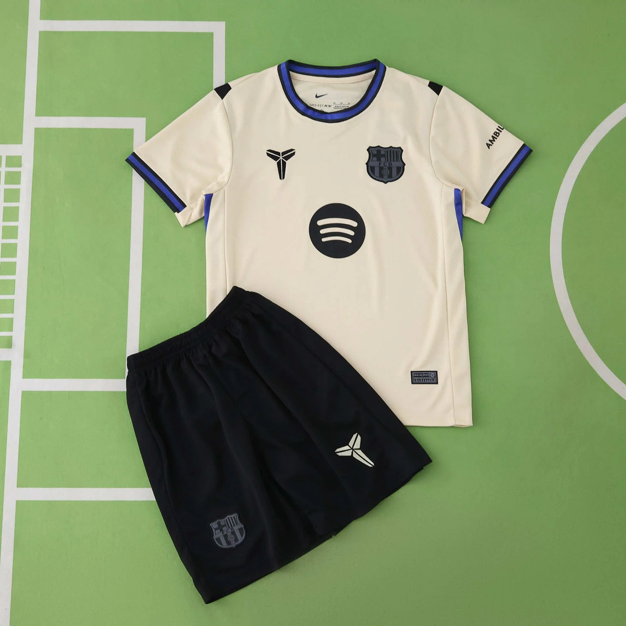 Soccer jersey and shorts laid out on a green sports court. The jersey is cream-colored with blue and black accents, featuring the FC Barcelona logo, a Spotify logo, and various sponsor logos. The black shorts have a FC Barcelona emblem and a logo on