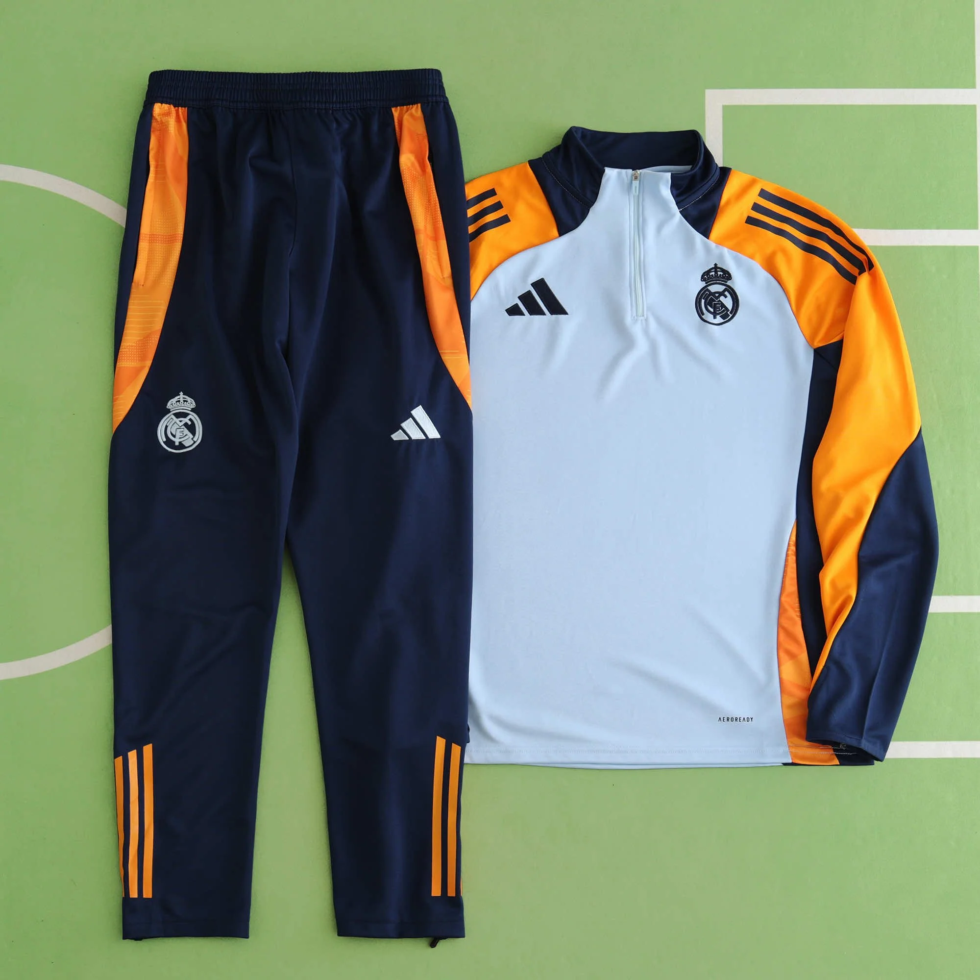 Real Madrid soccer training kit consisting of navy blue pants with orange accents and a white and orange training top, laid out on a green surface.