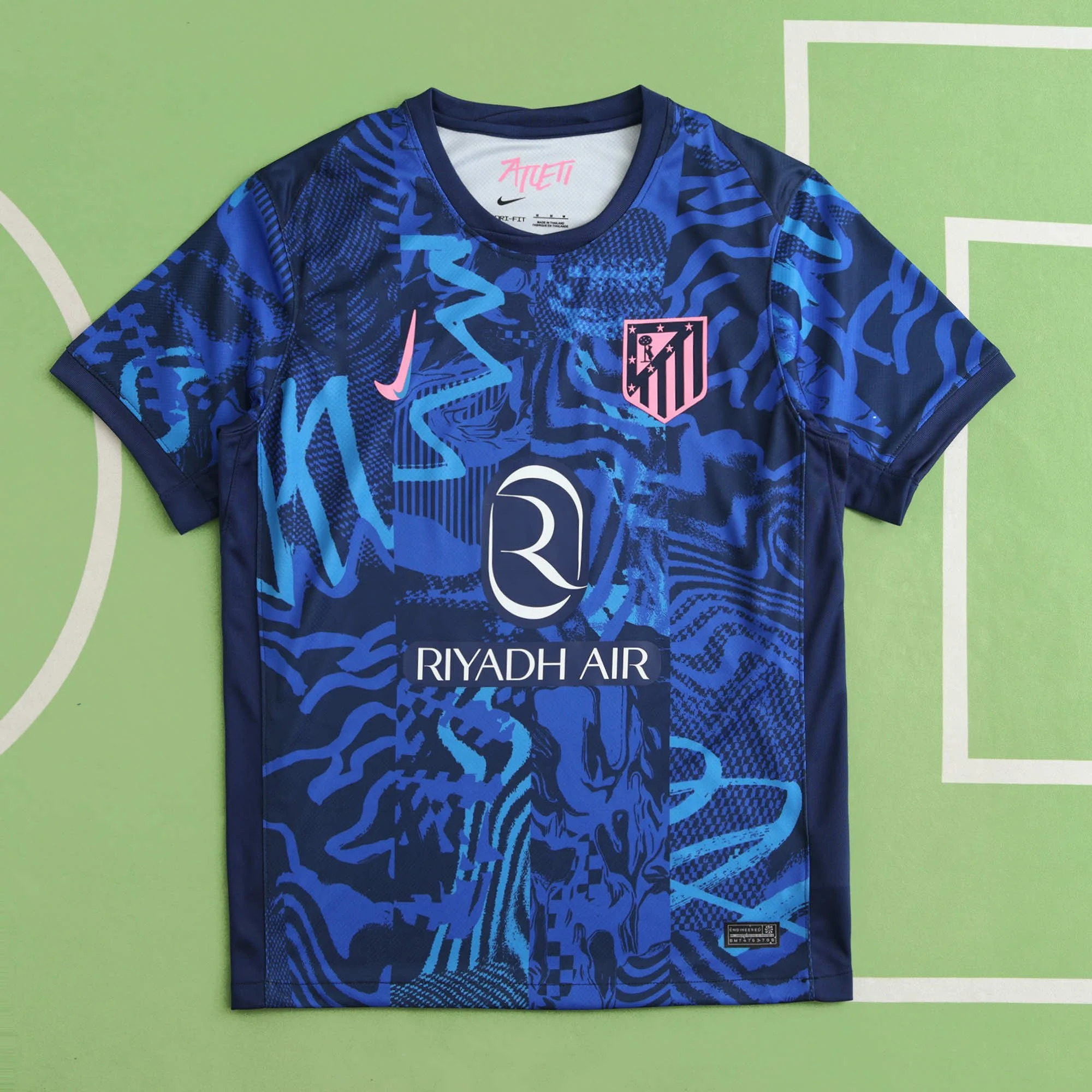 Blue sports jersey with abstract patterns and logos, including a pink Nike swoosh, a crest with black stripes and pink stars, and the words "Riyadh Air" on the front.