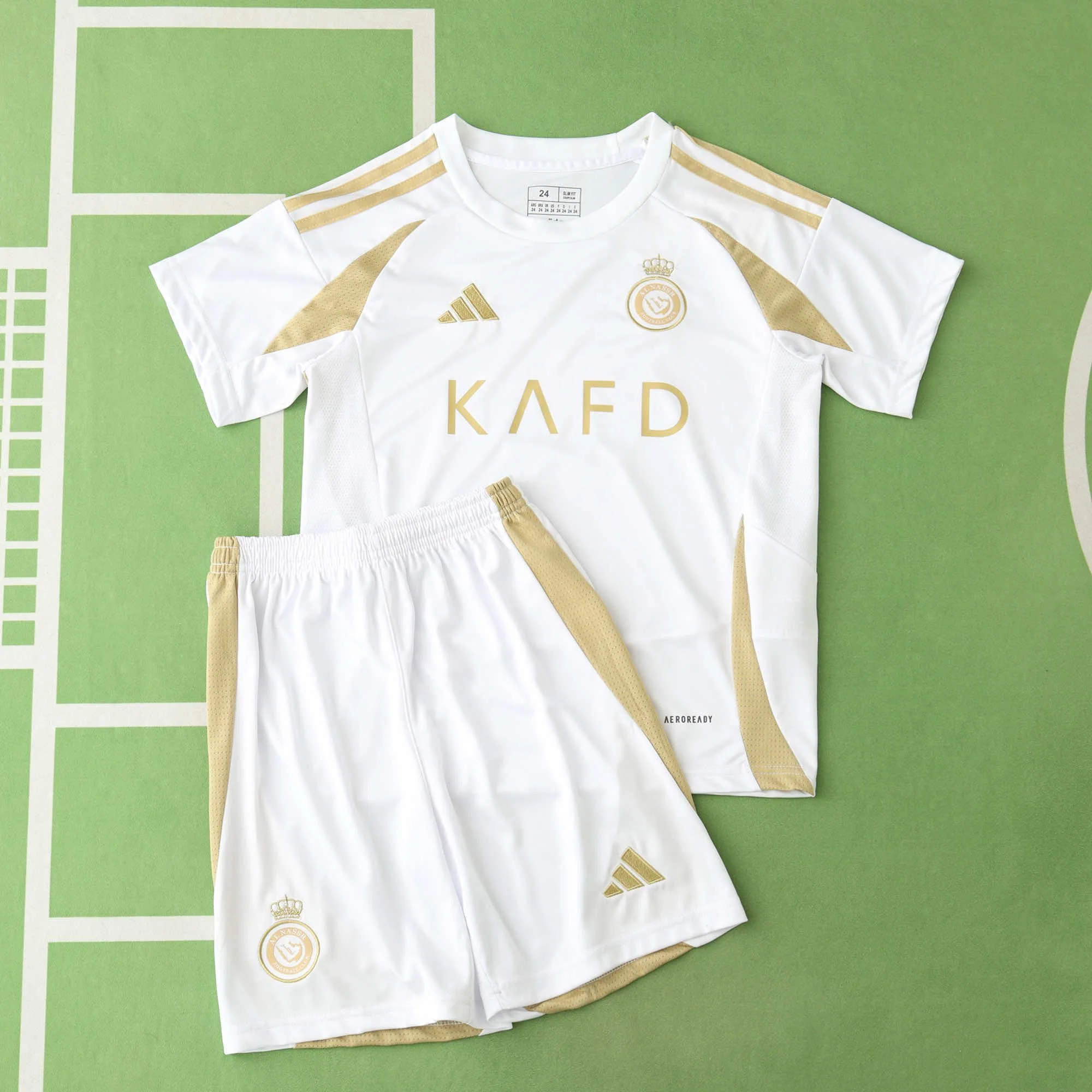 White sports jersey and shorts with gold accents displayed on a green surface, featuring Adidas logos and a crest with a crown.