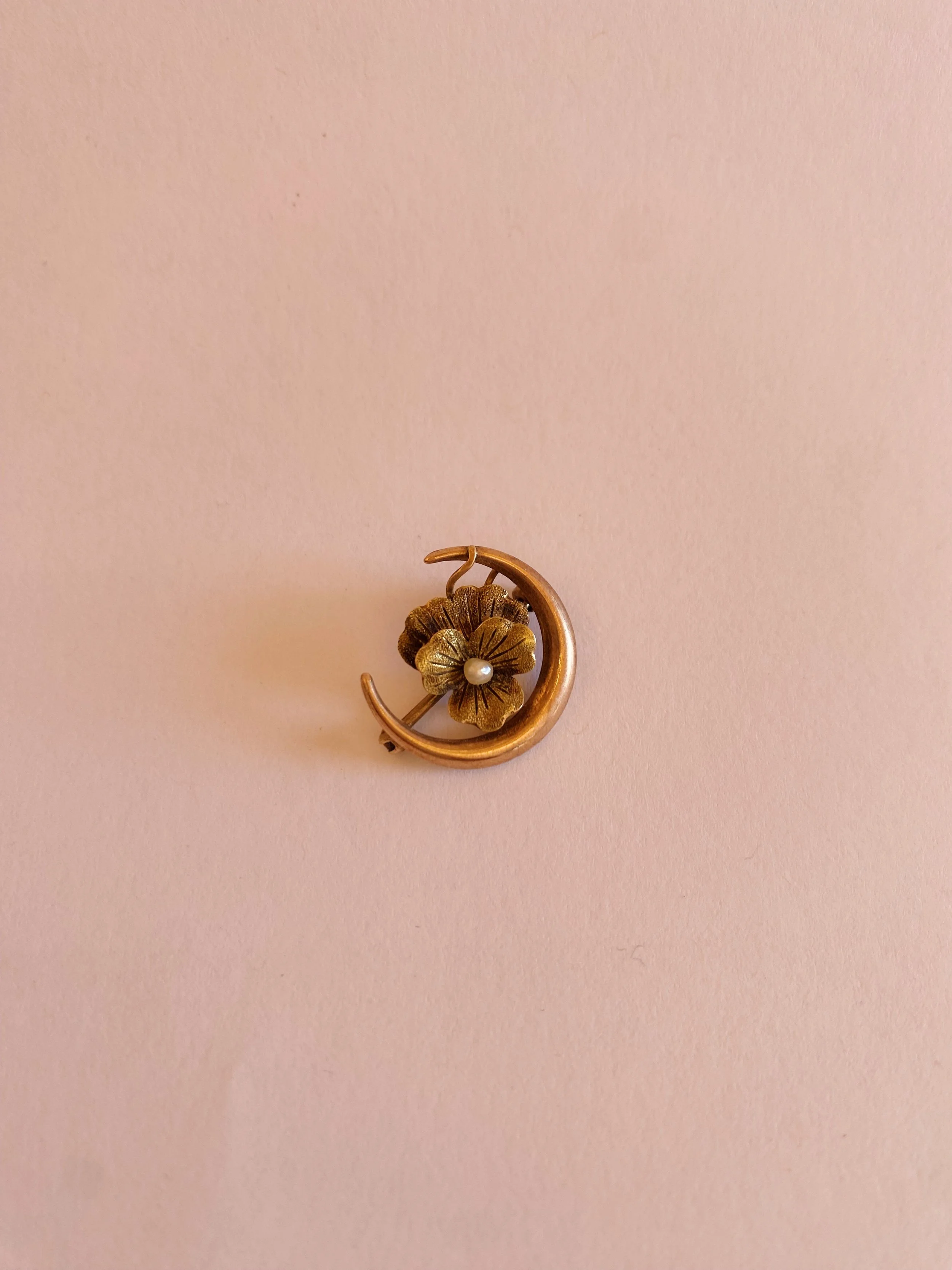A gold pin shaped like a flower with a pearl in the center, attached to a crescent moon, on a pink background.