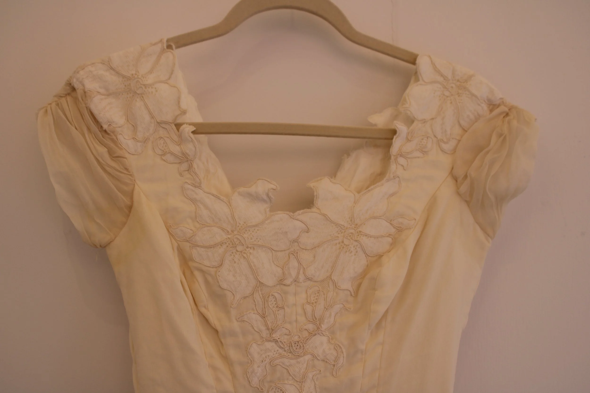 A cream-colored dress with floral embroidery hanging on a wooden hanger against a plain wall.