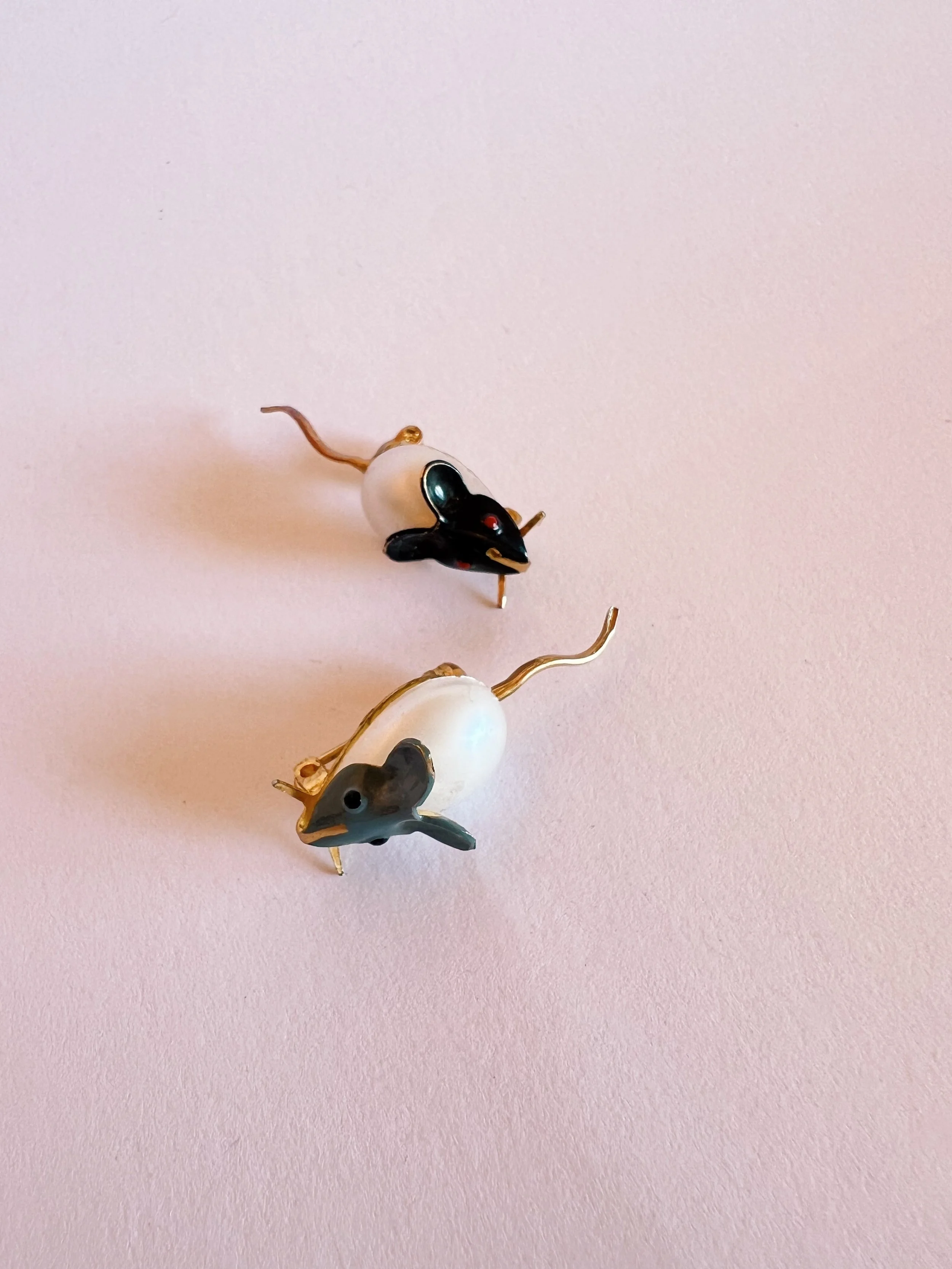 Two decorative mouse-shaped pins with black and white bodies and gold-colored tails, on a pink surface.