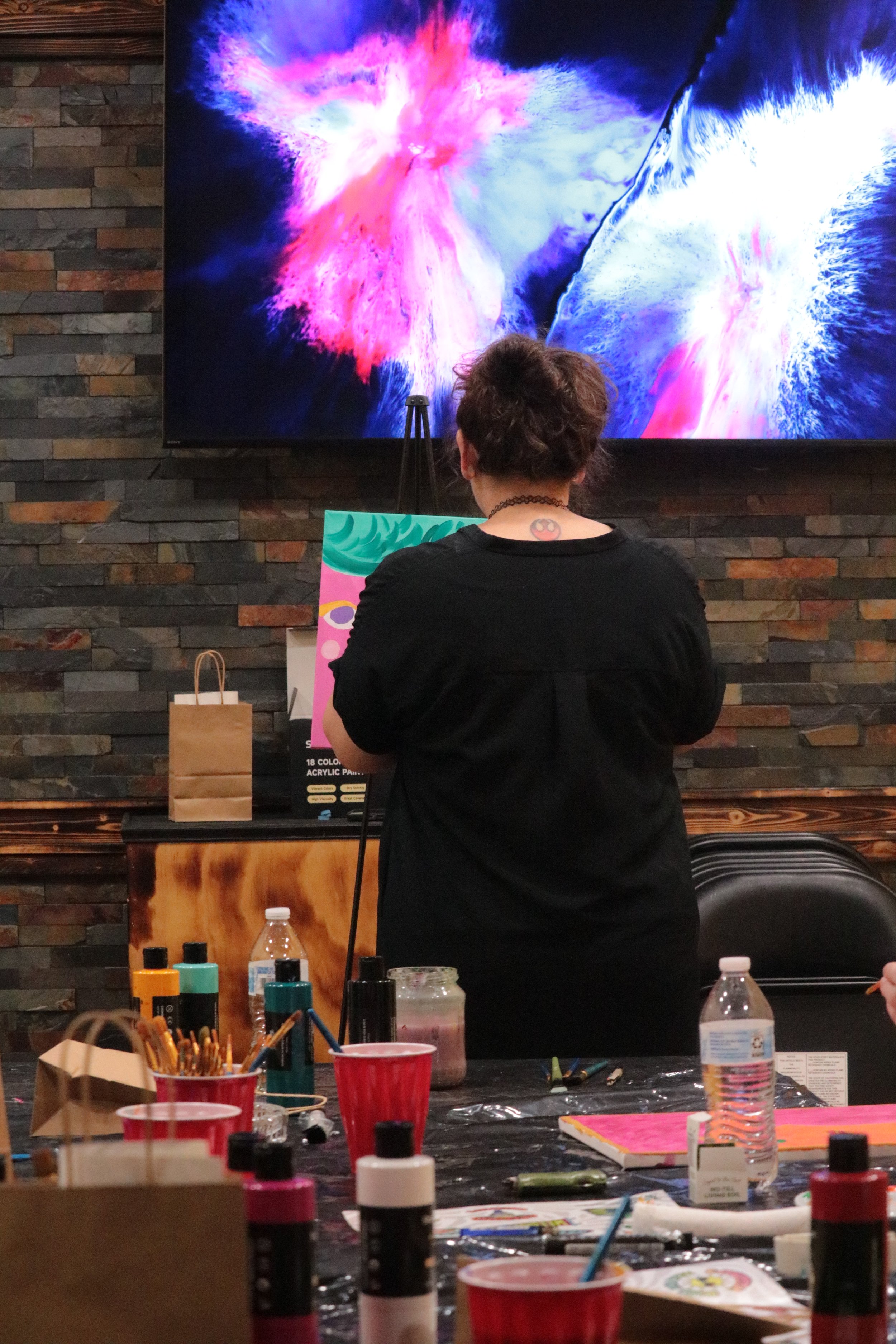A person with short brown hair, wearing a black shirt and choker, standing in front of a colorful abstract digital display featuring pink, blue, and white colors, in a room with a brick wall and a table covered with art supplies including paints, bru