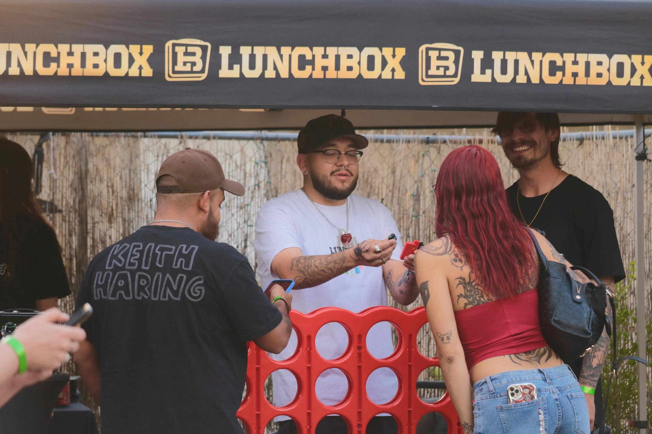 Group of people at an outdoor event, some with tattoos and colorful hair, gathered around a table under a canopy that has 'LUNCHBOX' written repeatedly. One person is smiling.