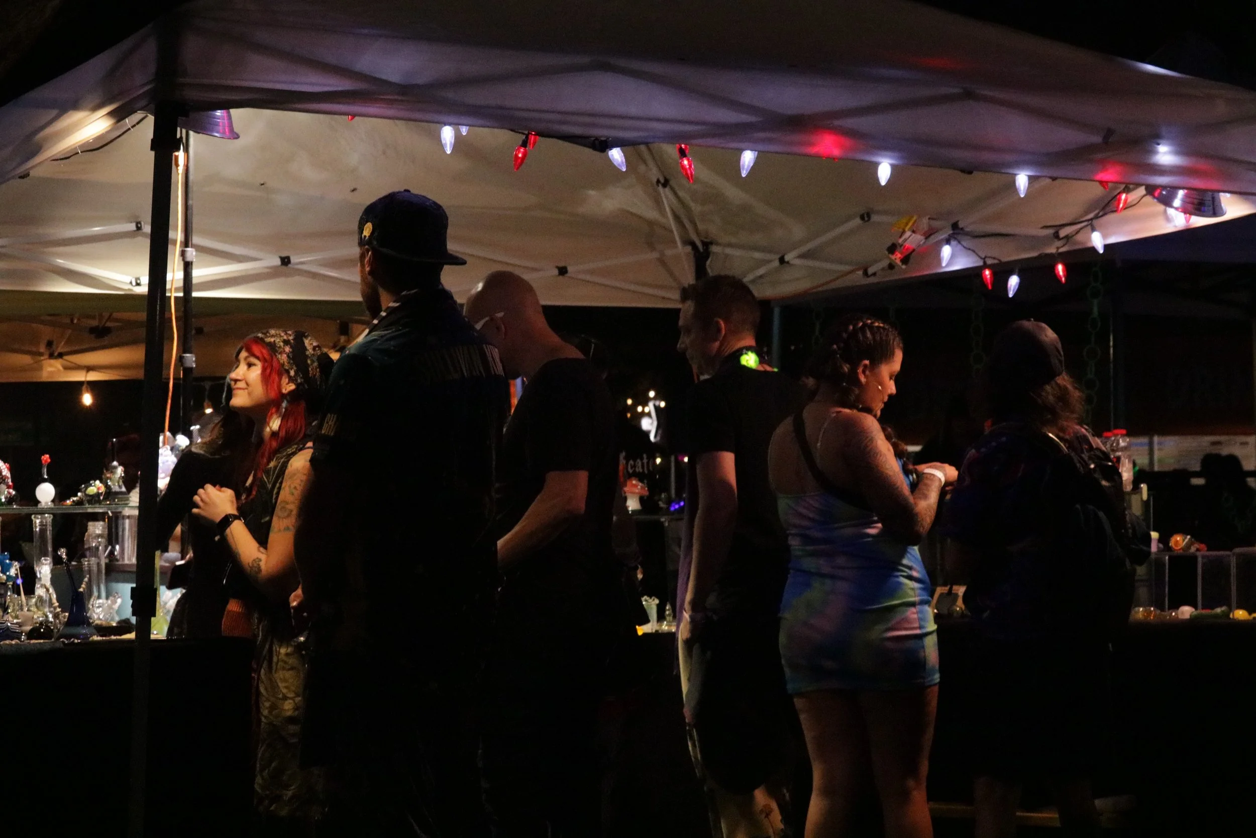 Nighttime outdoor scene with people gathered under a tent with string lights. A woman with red hair and tattoos is smiling, and others are engaged in conversation or looking at a display of glassware.