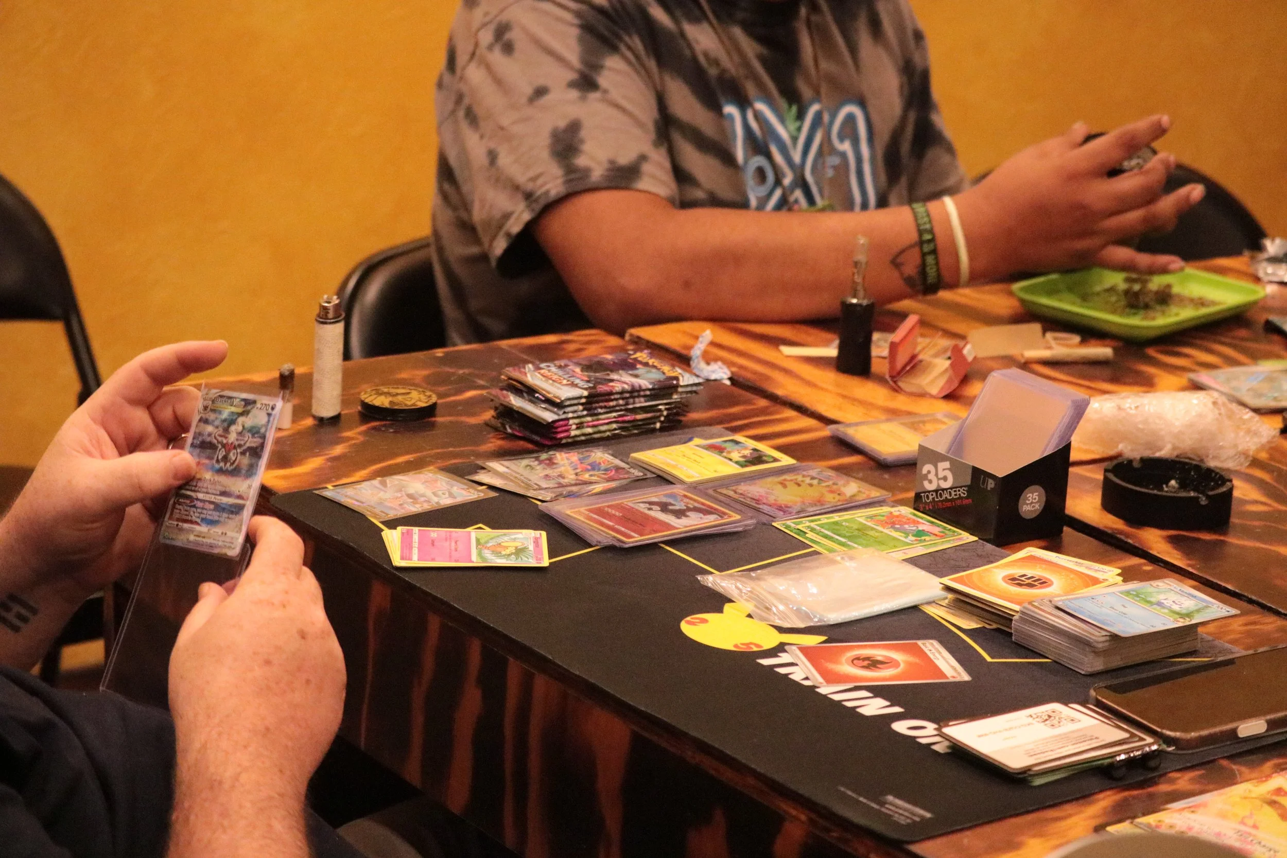 People playing a trading card game at a table with various cards, packs, and accessories, with a person holding a card in the foreground and another person sitting across with food on a green plate.