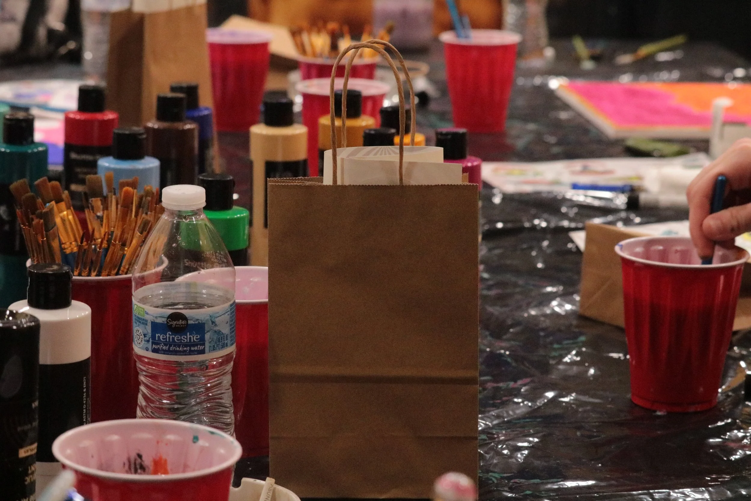 A table with art supplies, including bottles of paint, brushes, and cups, some filled with water, with the sponsored goodie bag of product samples