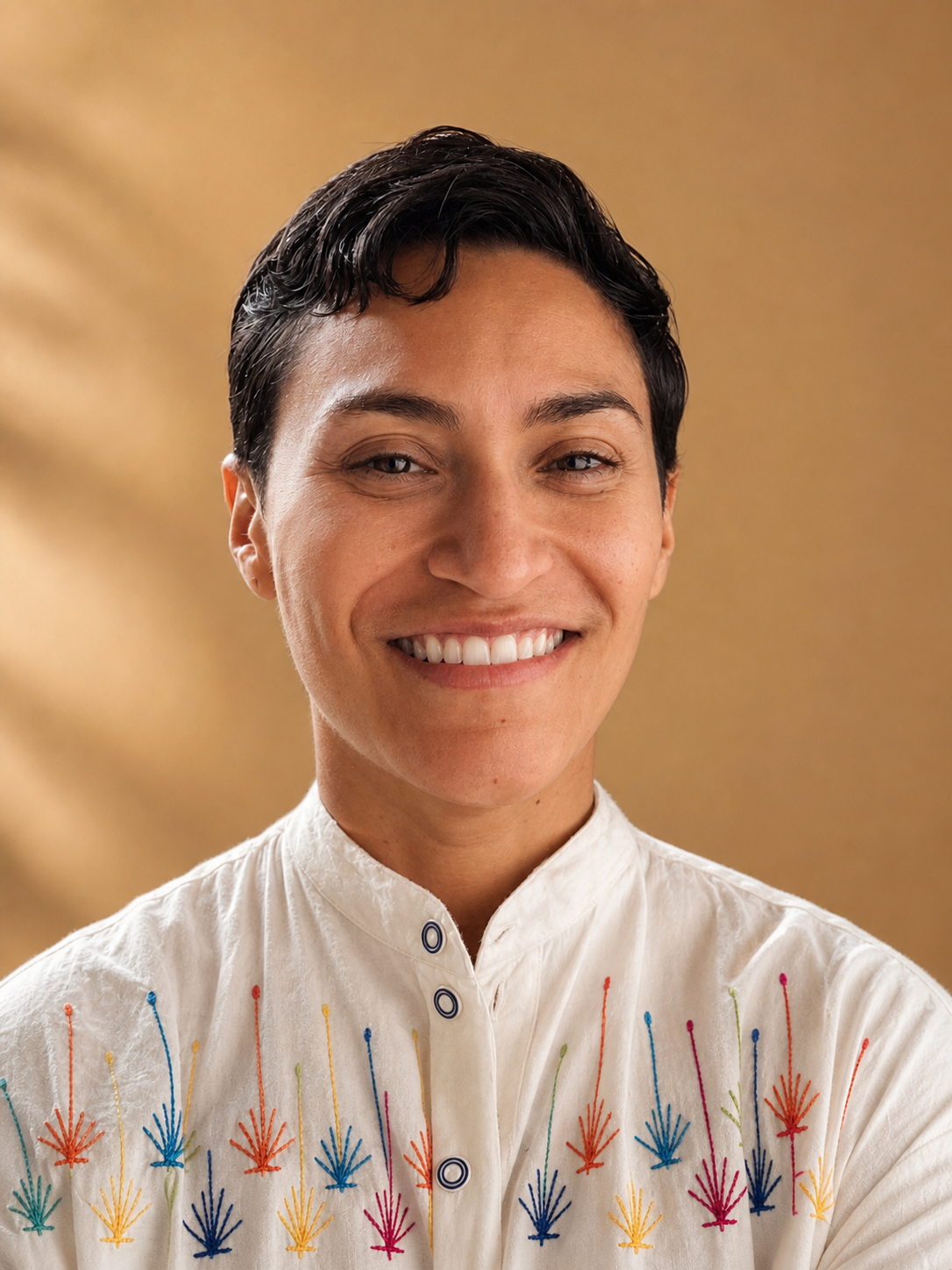 A smiling person with short dark hair wearing a white embroidered Mexican shirt against a warm background.