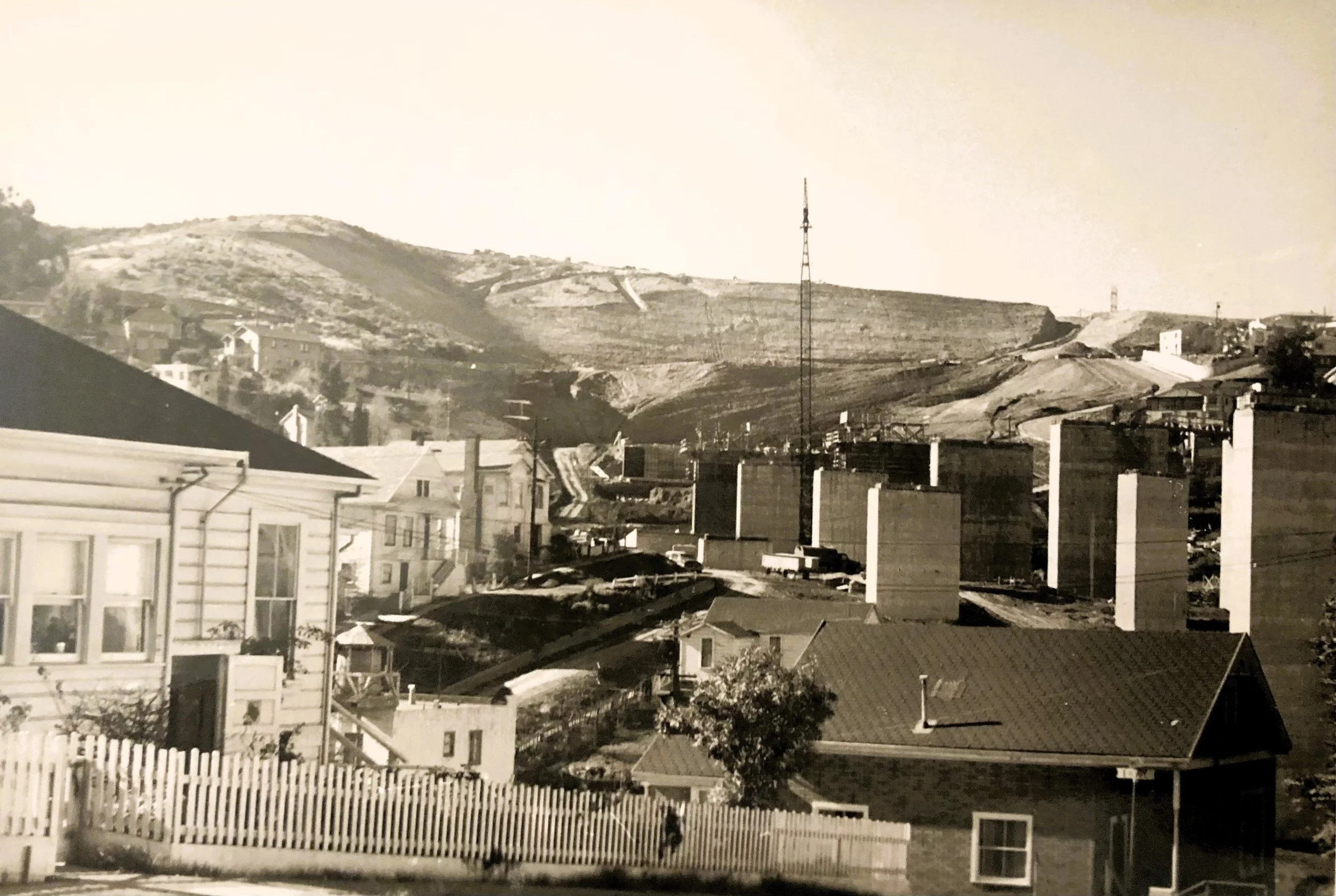 A black-and-white photo of a hillside neighborhood with various houses, a tall radio tower, and hilly terrain in the background.