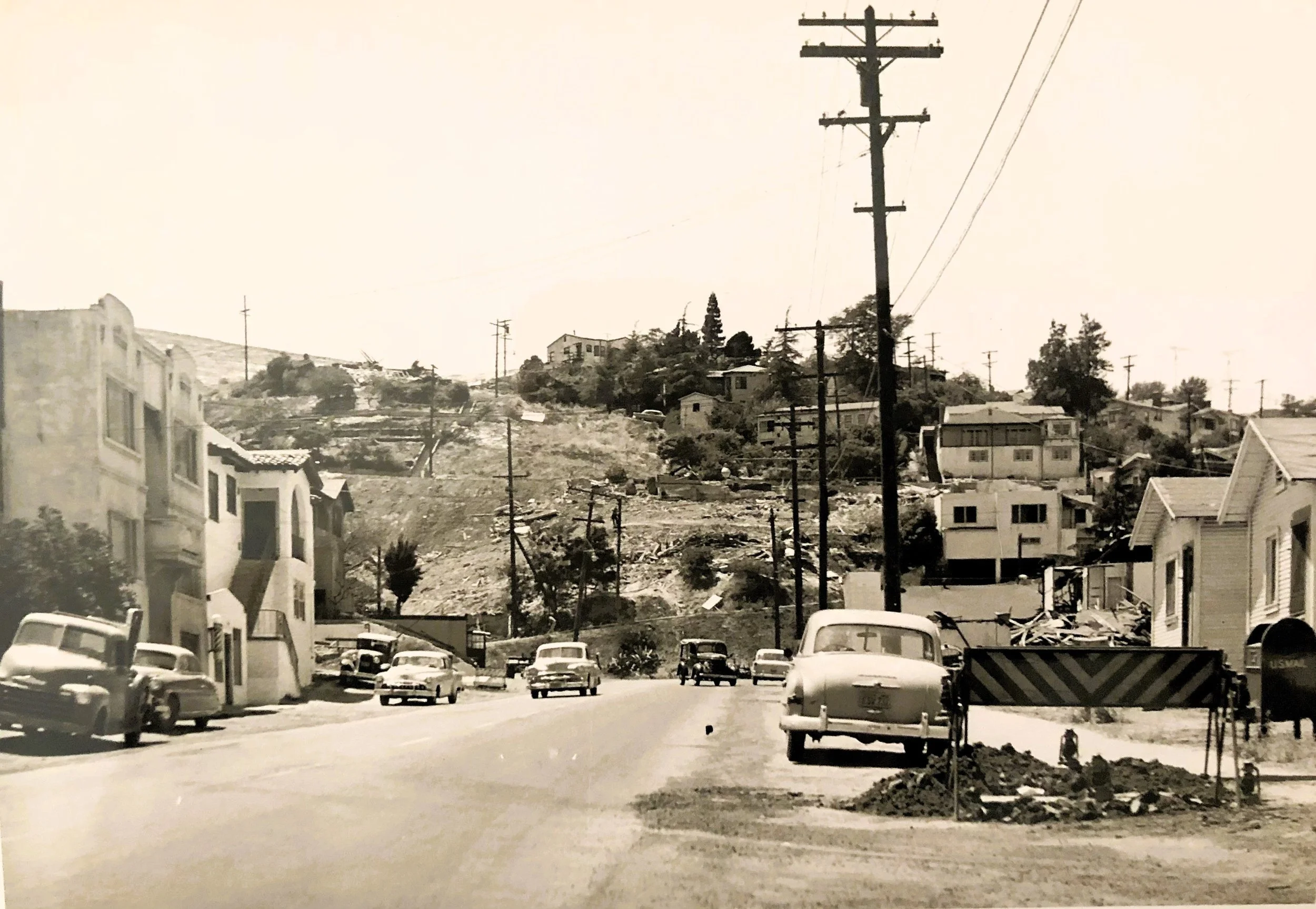 Black and white photo of a hilly residential neighborhood with vintage cars parked along the street, utility poles, and houses on both sides of the street and on the hill.