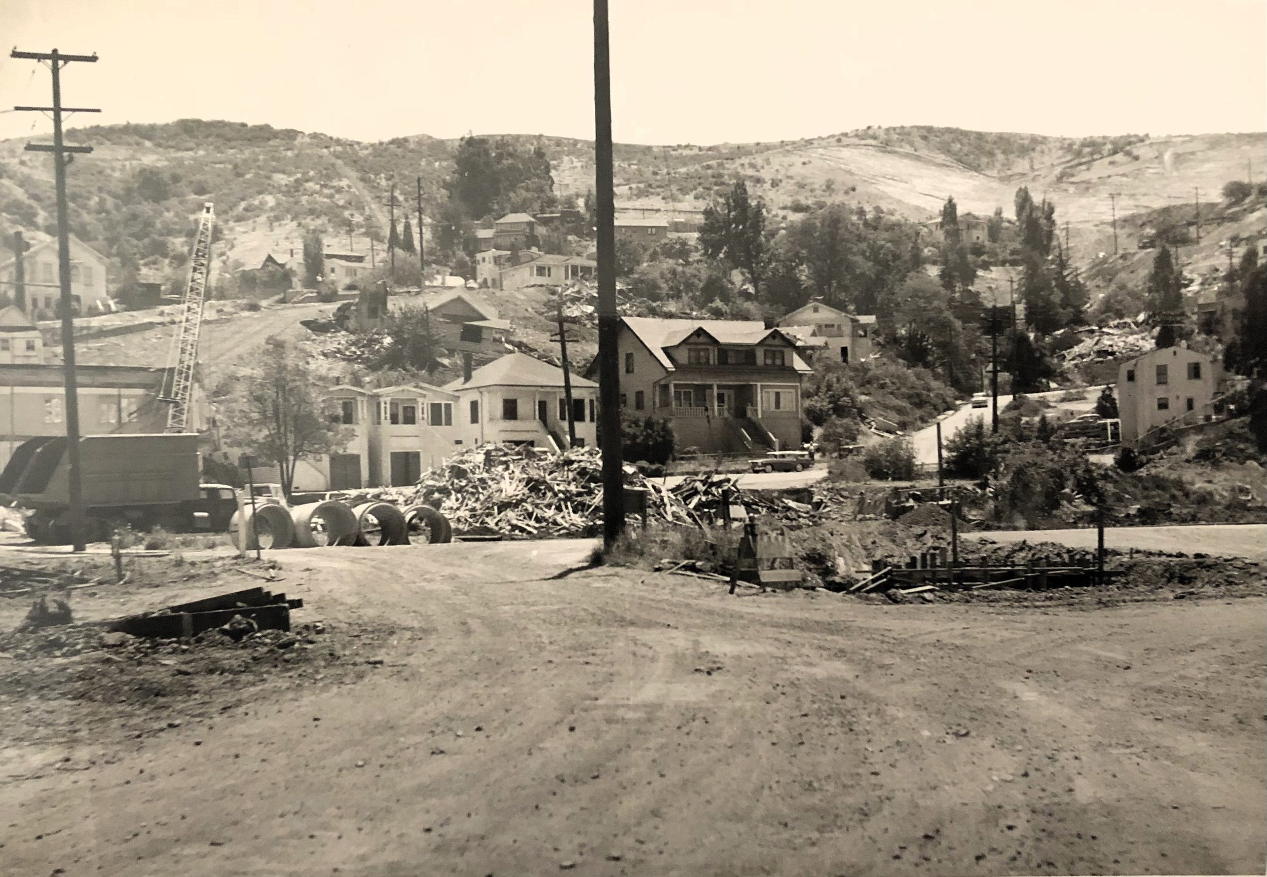 A black and white photo of a hilly residential area with houses, dirt roads, construction debris, and utility poles.