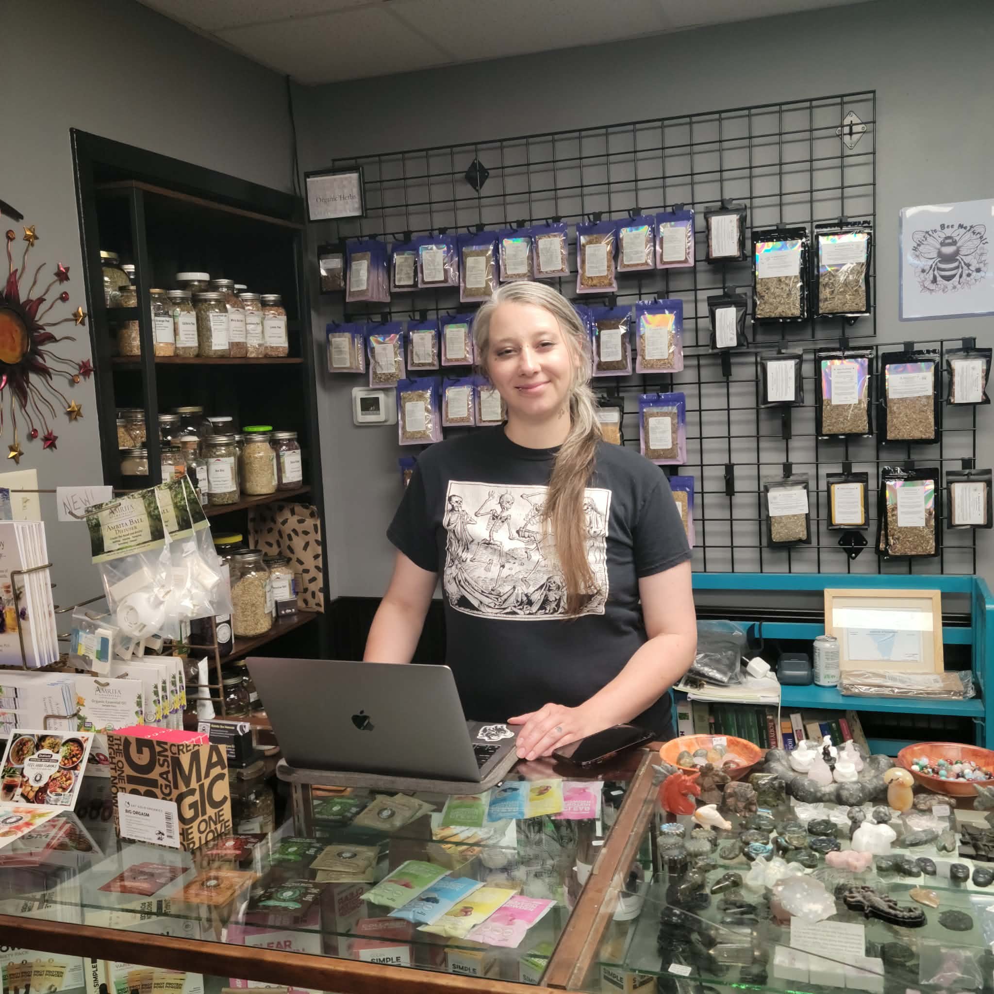 Brycee stands before her laptop, smiling for the camera behind behind her retail corner inside Obsidian & Sage, Moline, IL 