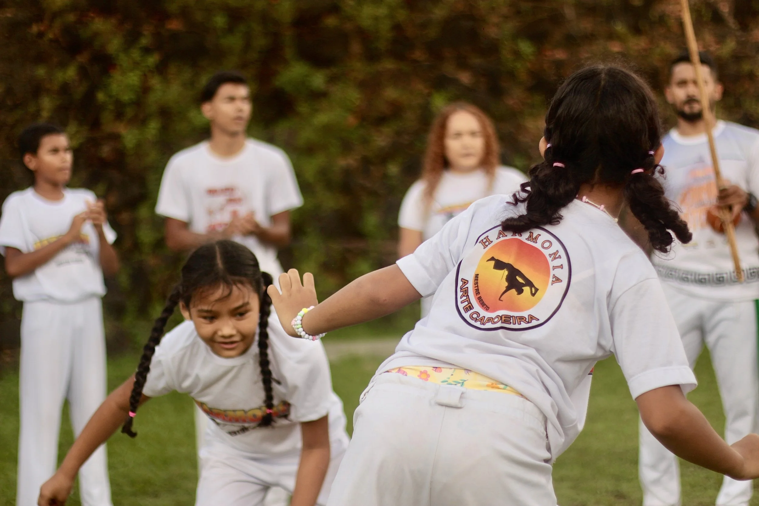 Crianças praticando capoeira ao ar livre, vestindo uniformes brancos. A menina na frente está com cabelo em duas tranças e se ajoelha enquanto uma garota com cabelo cacheado observa ao fundo, com uma roda de crianças ao redor.