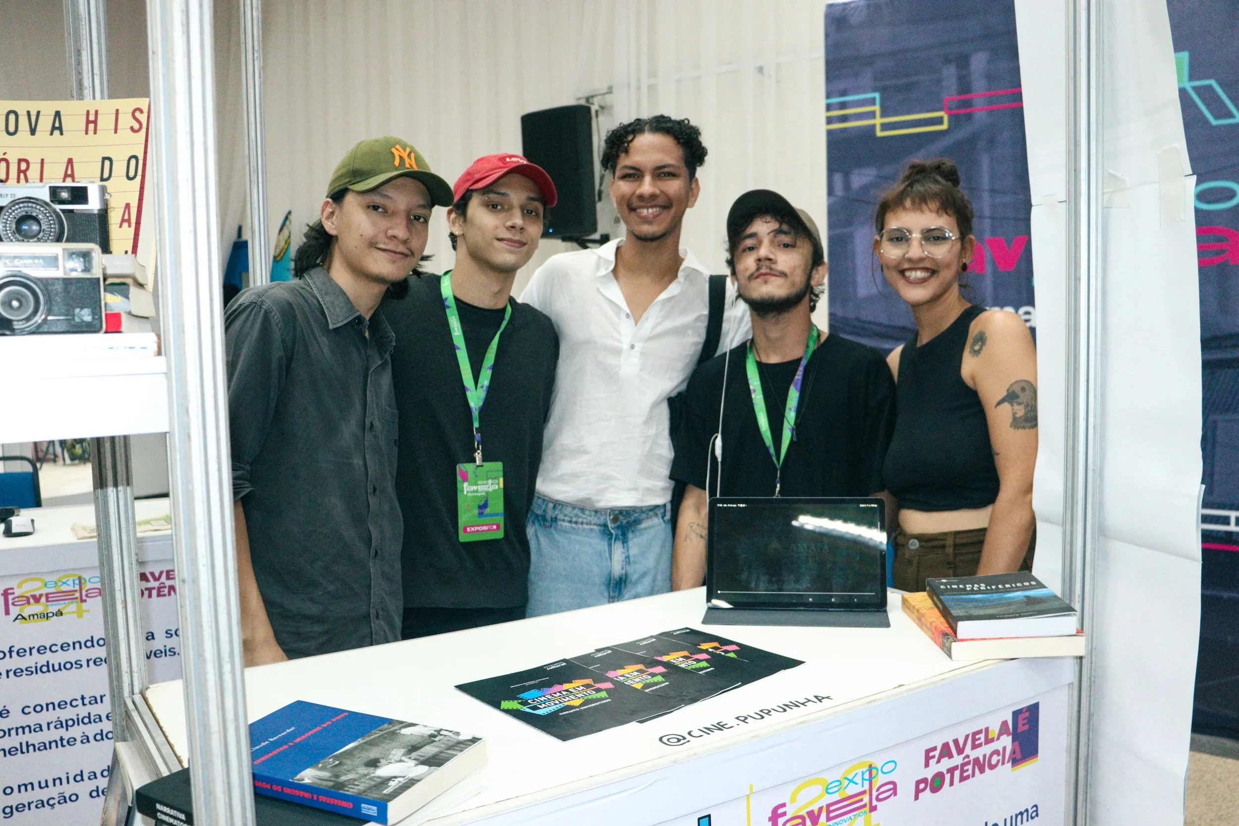 Grupo de cinco jovens sorrindo em estande de feira, com banner e livros na mesa.