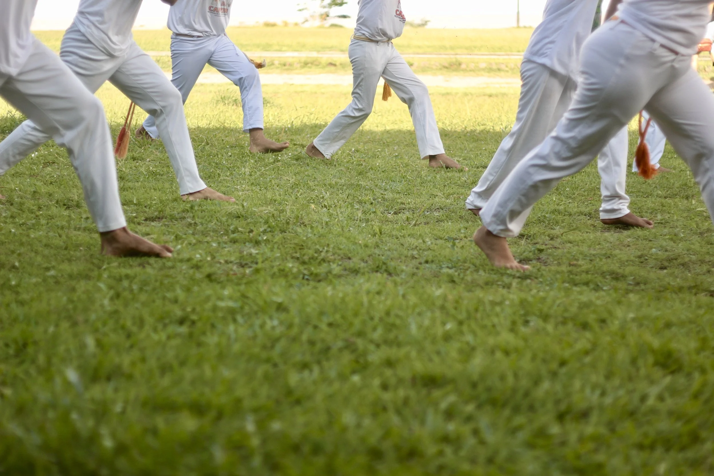 Praticantes de capoeira treinando ao ar livre, vestindo roupas brancas tradicionais e descalços, em um campo de grama.