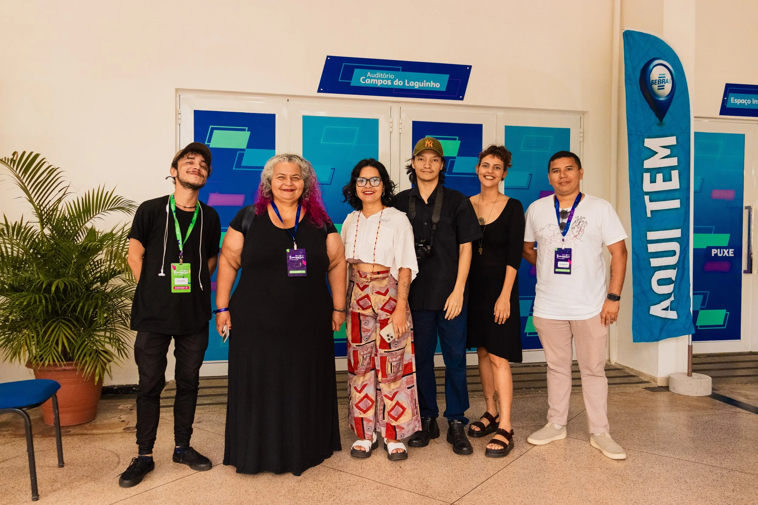 Grupo de seis pessoas posando para foto em evento, em frente a uma porta com sinalização azul, com uma bandeira azul ao lado esquerdo e plantas na lateral esquerda.