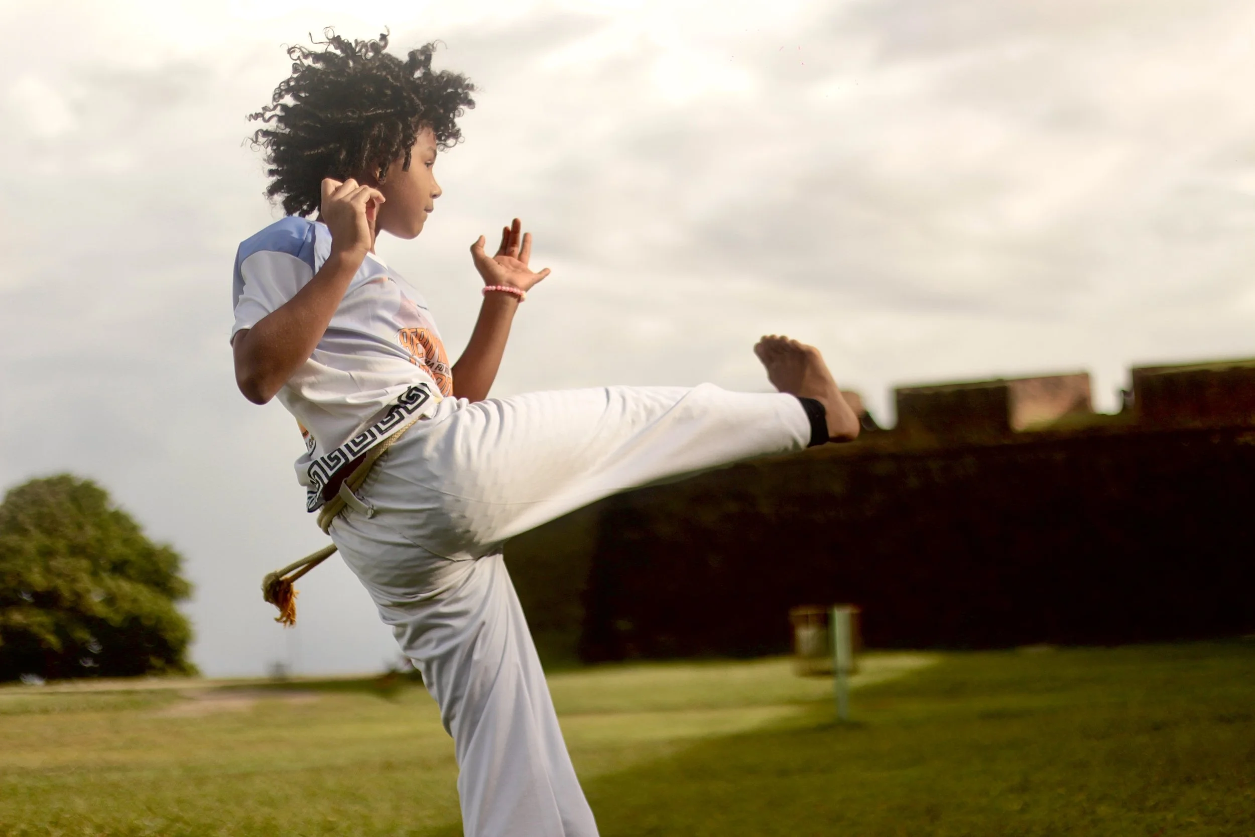 Pessoa praticando capoeira, executando um golpe de destaque, ao ar livre com céu nublado ao fundo.