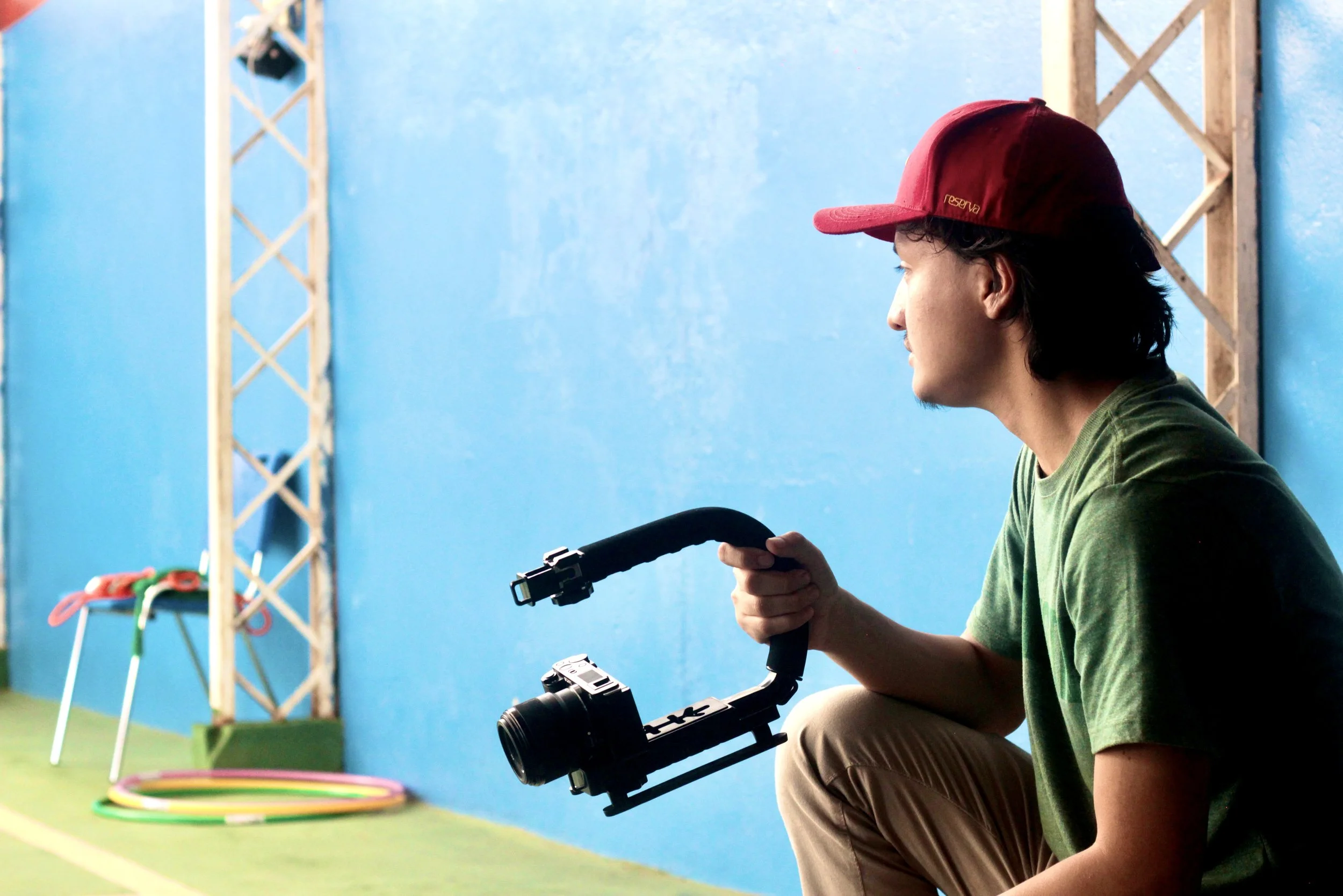 Homem sentado com câmera gimbal na mão, assistindo em um espaço com parede azul, equipamentos de suporte de ginástica ao fundo.