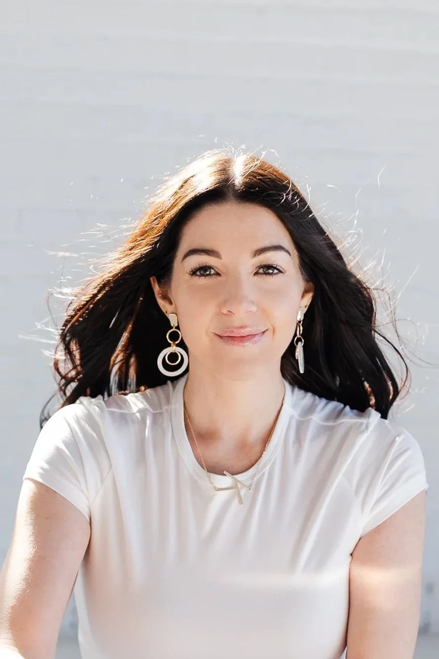 A woman with long dark hair wearing a white shirt and large earrings, smiling outdoors against a bright background.