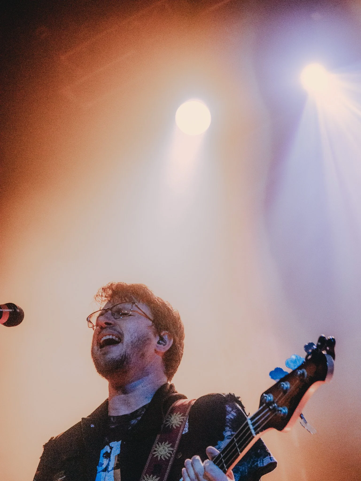 A musician with glasses and a floral shirt playing an electric guitar on stage, illuminated by stage lights.