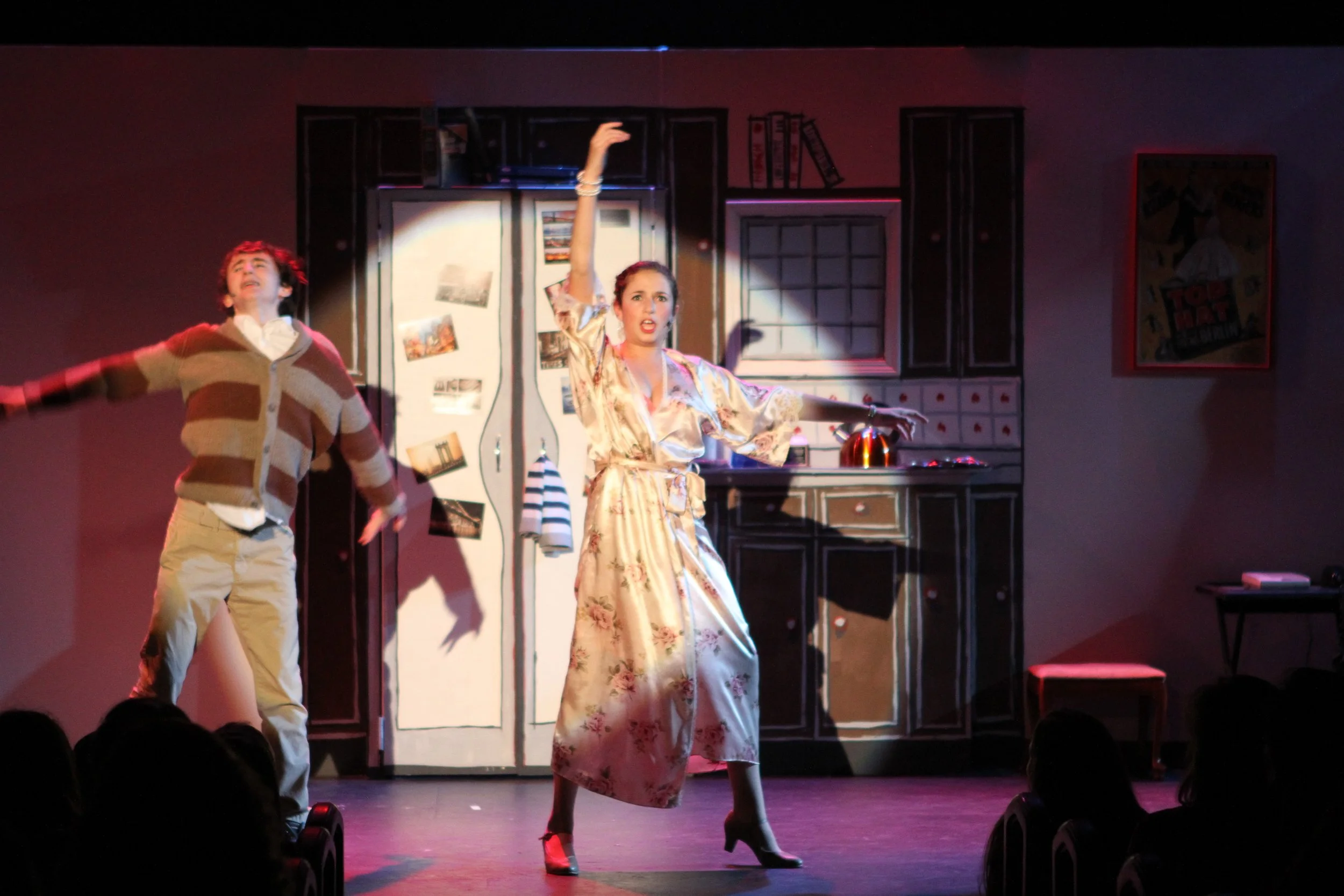 Two actors performing on stage in a theatrical production, with a kitchen set in the background.