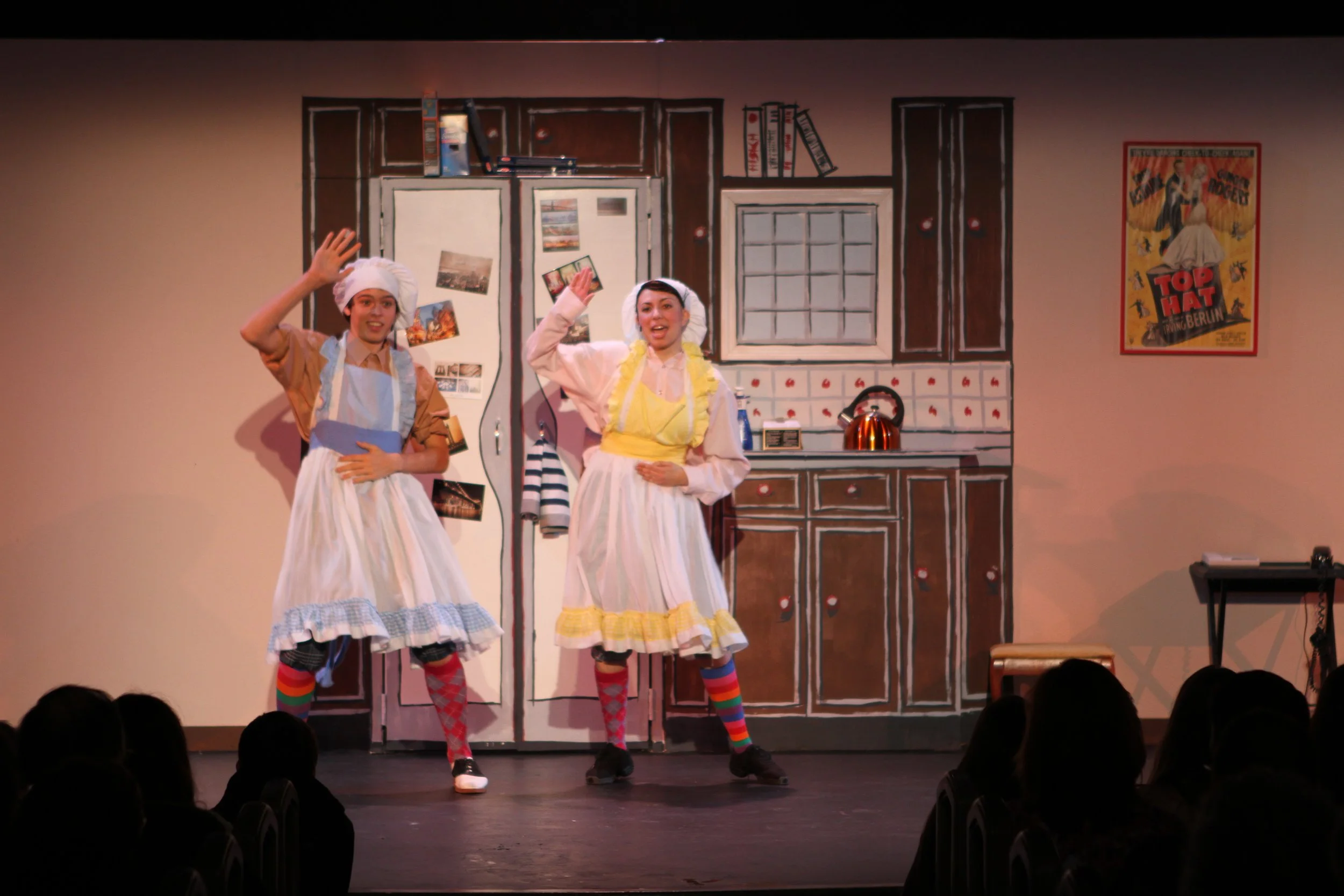 The image shows two young women with expressive faces performing on stage, dressed in costumes with aprons, head coverings, and striped socks, in a kitchen set with painted cabinets,, and a poster in the background, audience visible at the bottom.