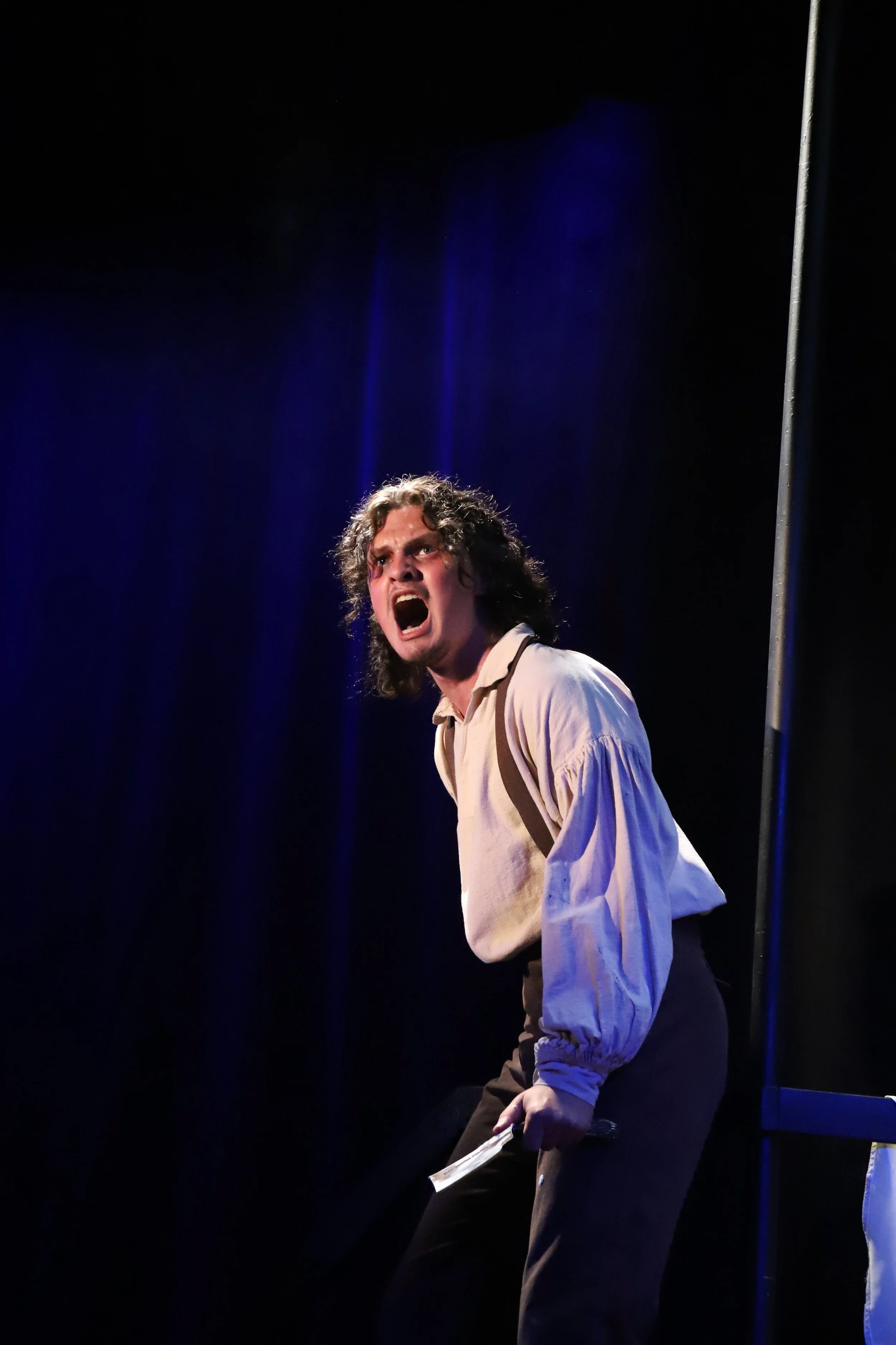 A person with curly hair, wearing a loose beige shirt and dark pants, appears to be performing on stage with an intense and emotional expression, illuminated by stage lights against a dark background.