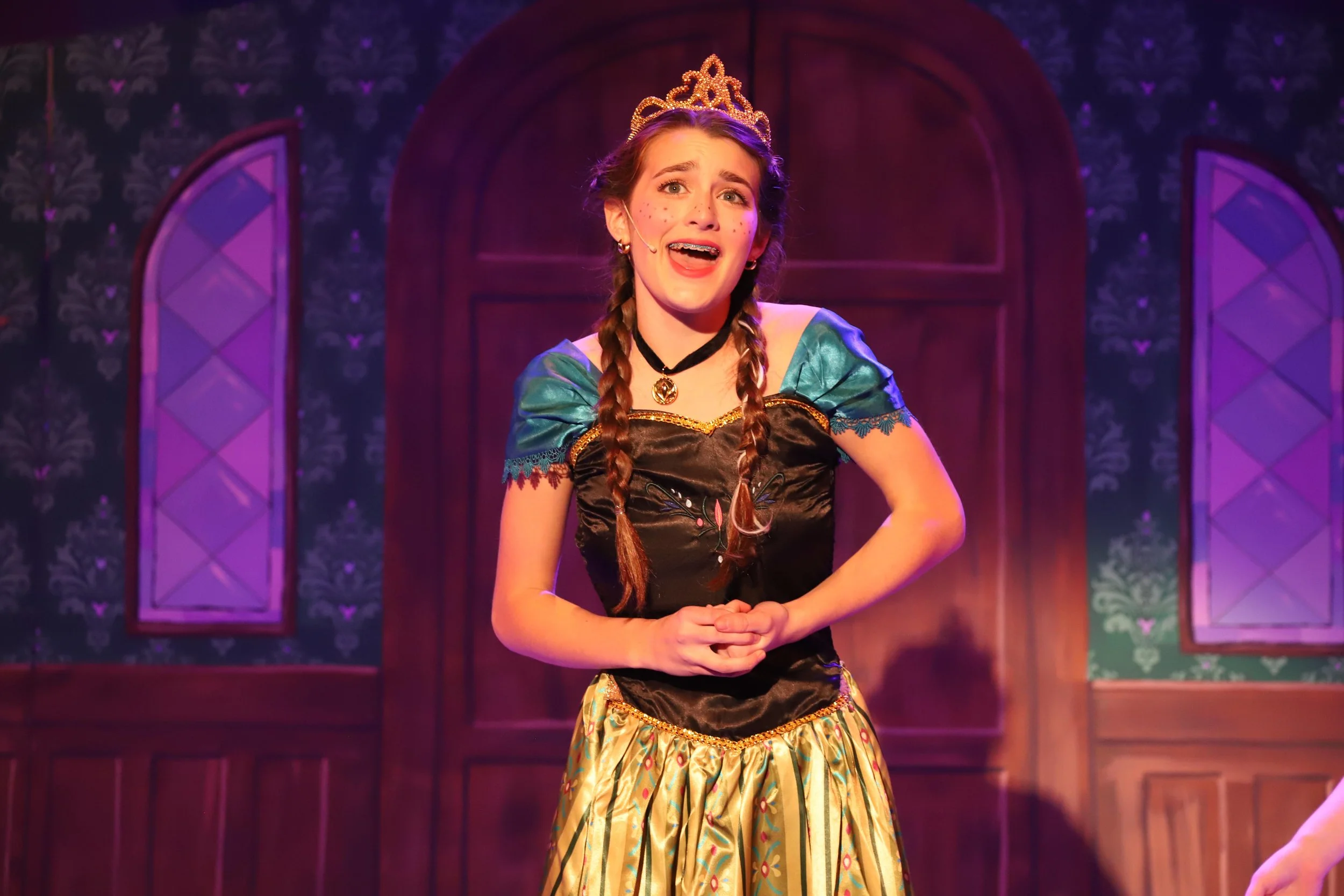 Young woman dressed as Anna in Frozen Jr. with a tiara, long braided hair, and wearing a colorful dress, appearing distressed or emotional, on stage with a purple and blue background.