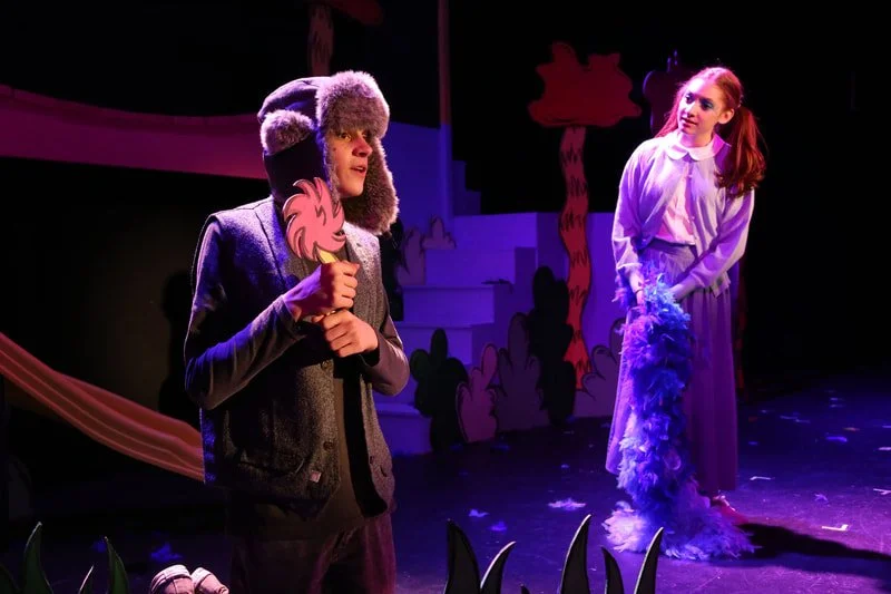 Two young actors perform on stage in a colorful theatrical play. The boy on the left is holding a pink and orange prop and wearing a furry hat with ear flaps, a jacket, and pants. The girl on the right wears a long skirt, a blouse, and is holding a l