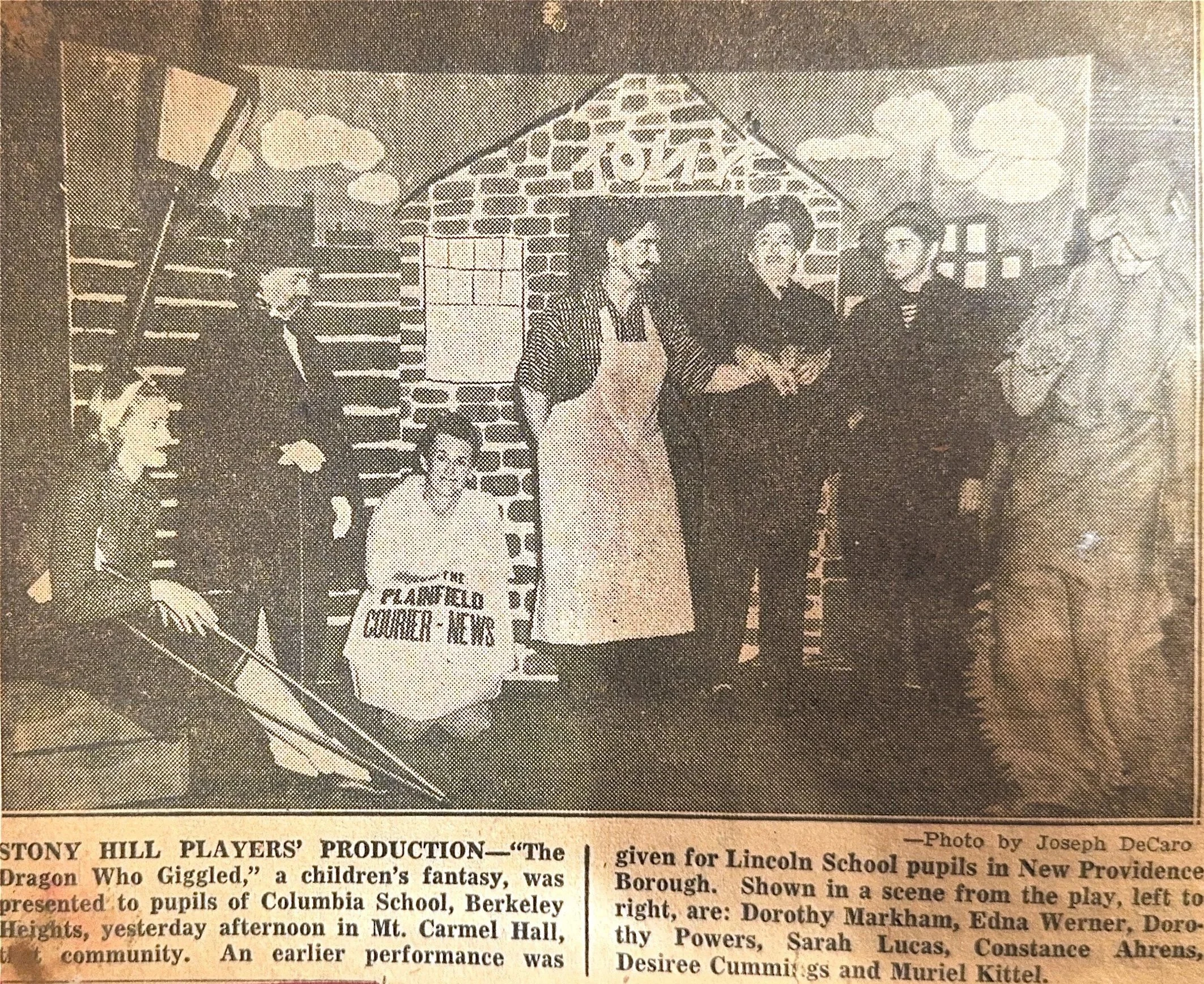 Children and adults performing in a children's fantasy play titled 'The Dragon Who Giggled' in a school auditorium with a brick backdrop. Several are holding scripts or props, including a small girl holding a sign.