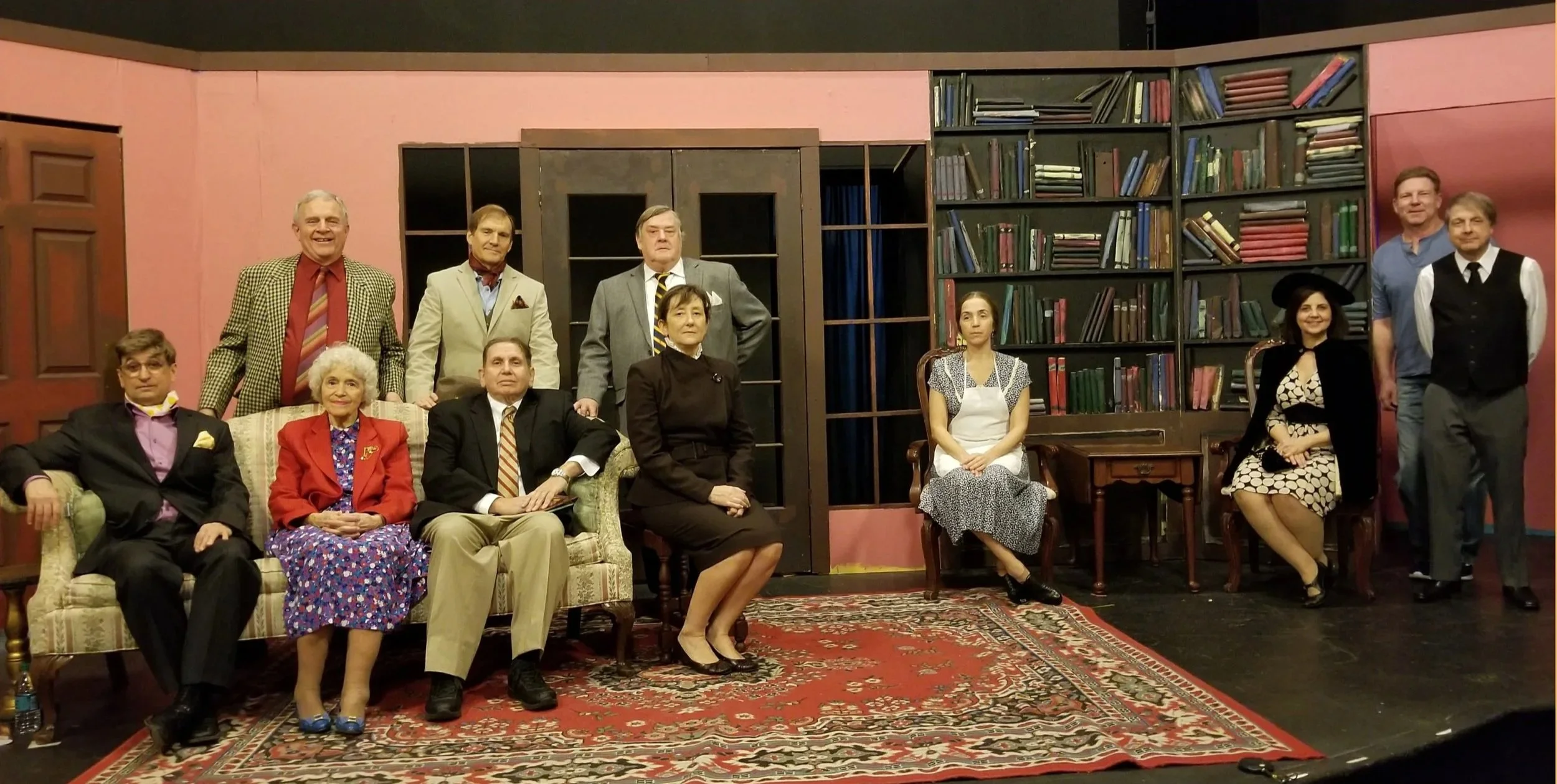 Group of twelve actors on a stage, arranged for a theatrical scene, with a background of bookshelves and pink walls, dressed in vintage costumes.