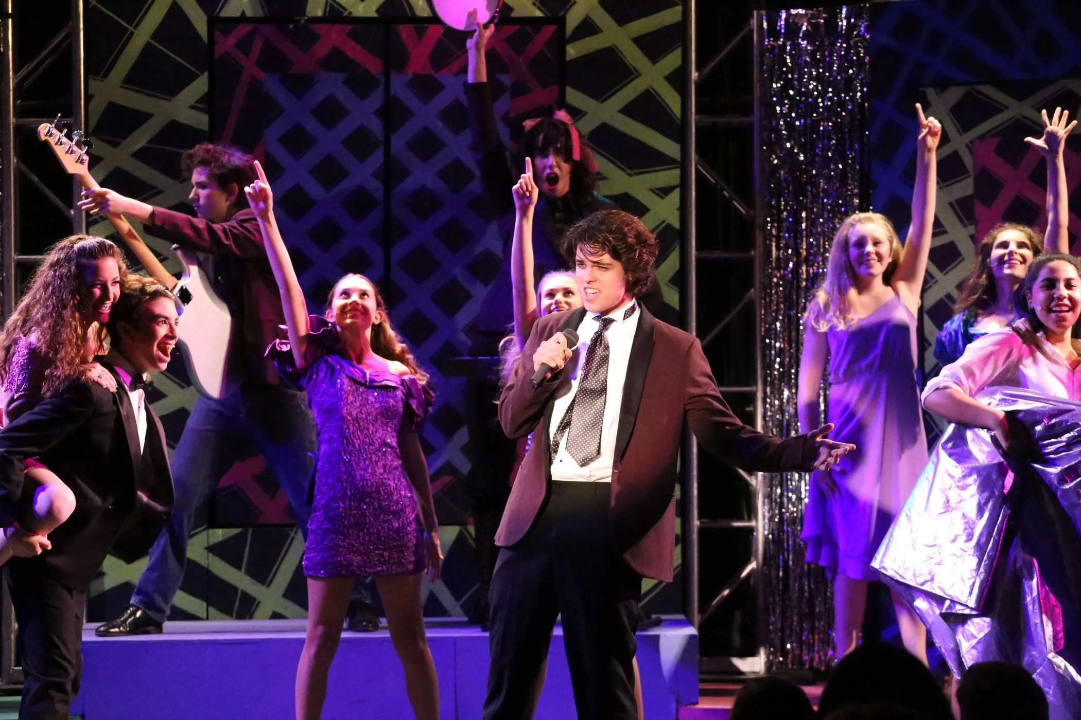 A theatrical stage scene with a group of young performers, some holding microphones and others playing instruments, dressed in colorful costumes, with vibrant lighting and a decorative backdrop.