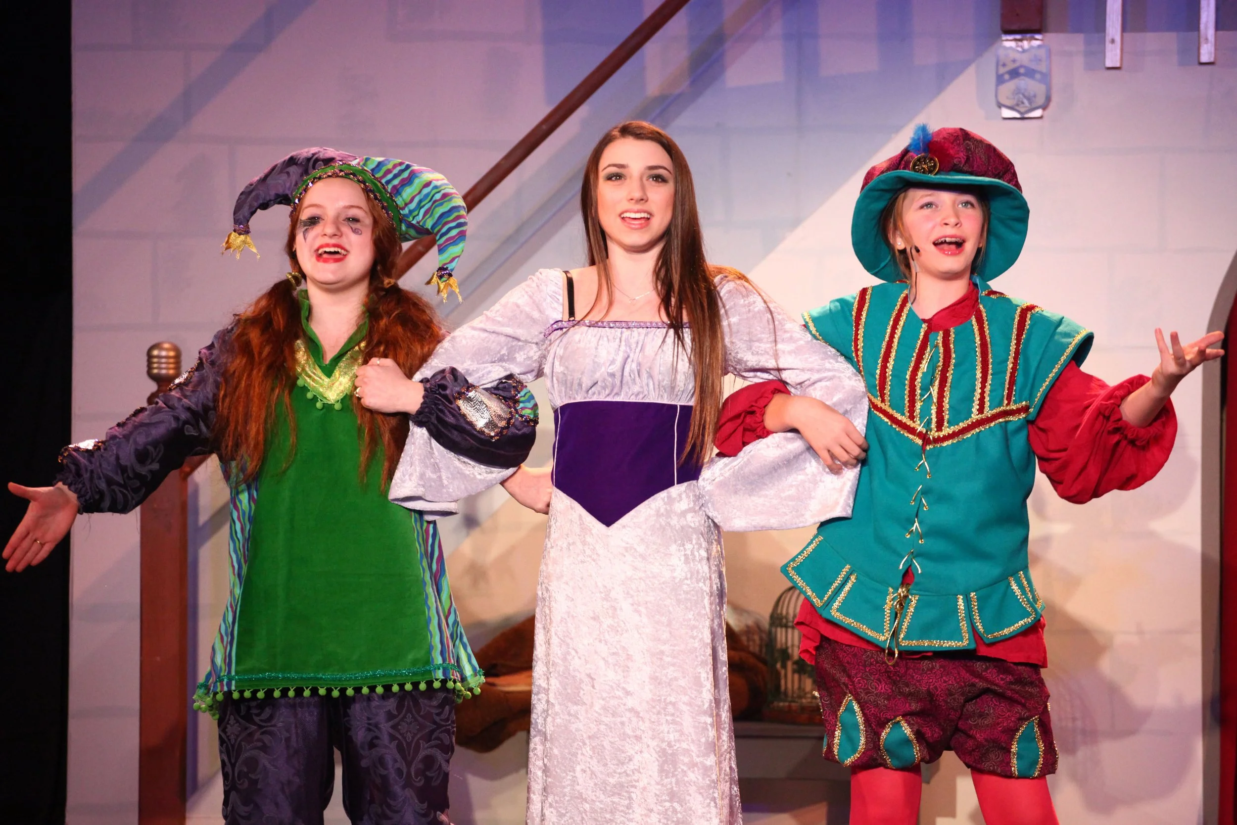 Three actresses performing on stage in colorful costumes. The middle actress, dressed in a princess gown, holds hands with the actresses on either side who are dressed as jesters. The background features a staircase with a coat of arms.
